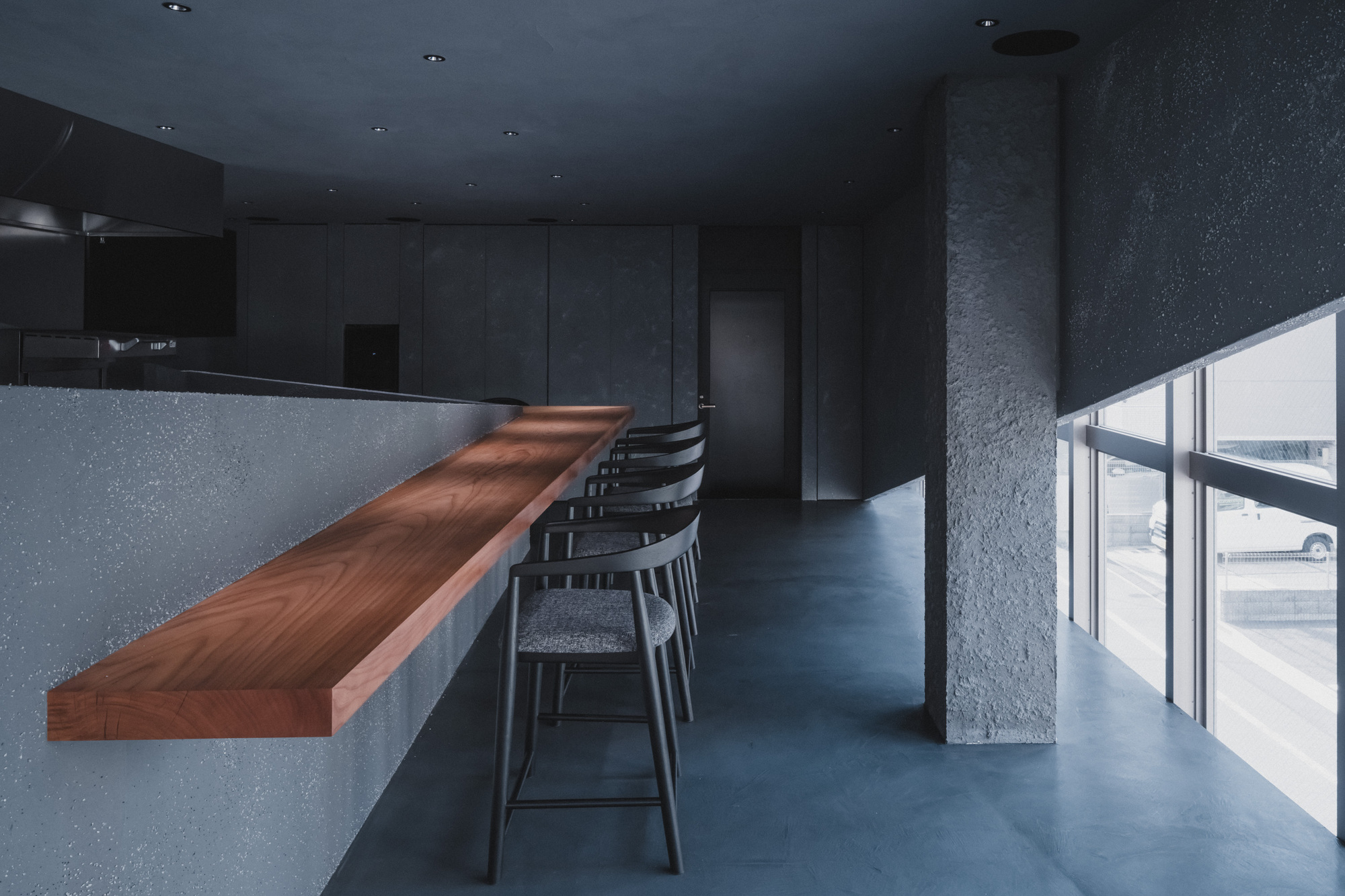 Cantilevered wood counter and stools beside a textured concrete column, with a parked car visible through the window glazing
