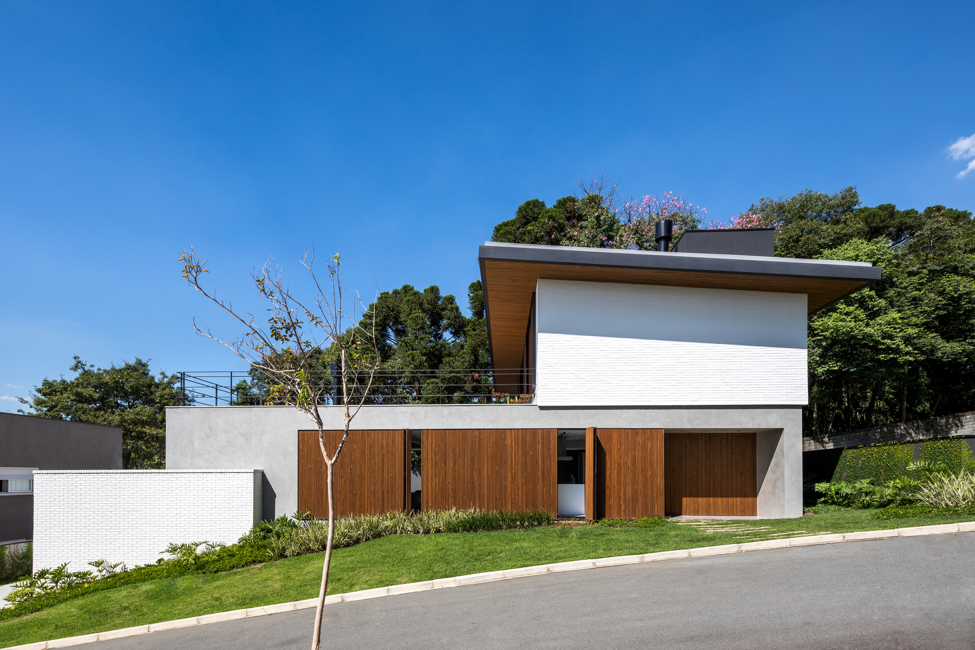 Street facade with sliding timber panels beneath a white brick upper volume and cantilevered roof, under a clear blue sky