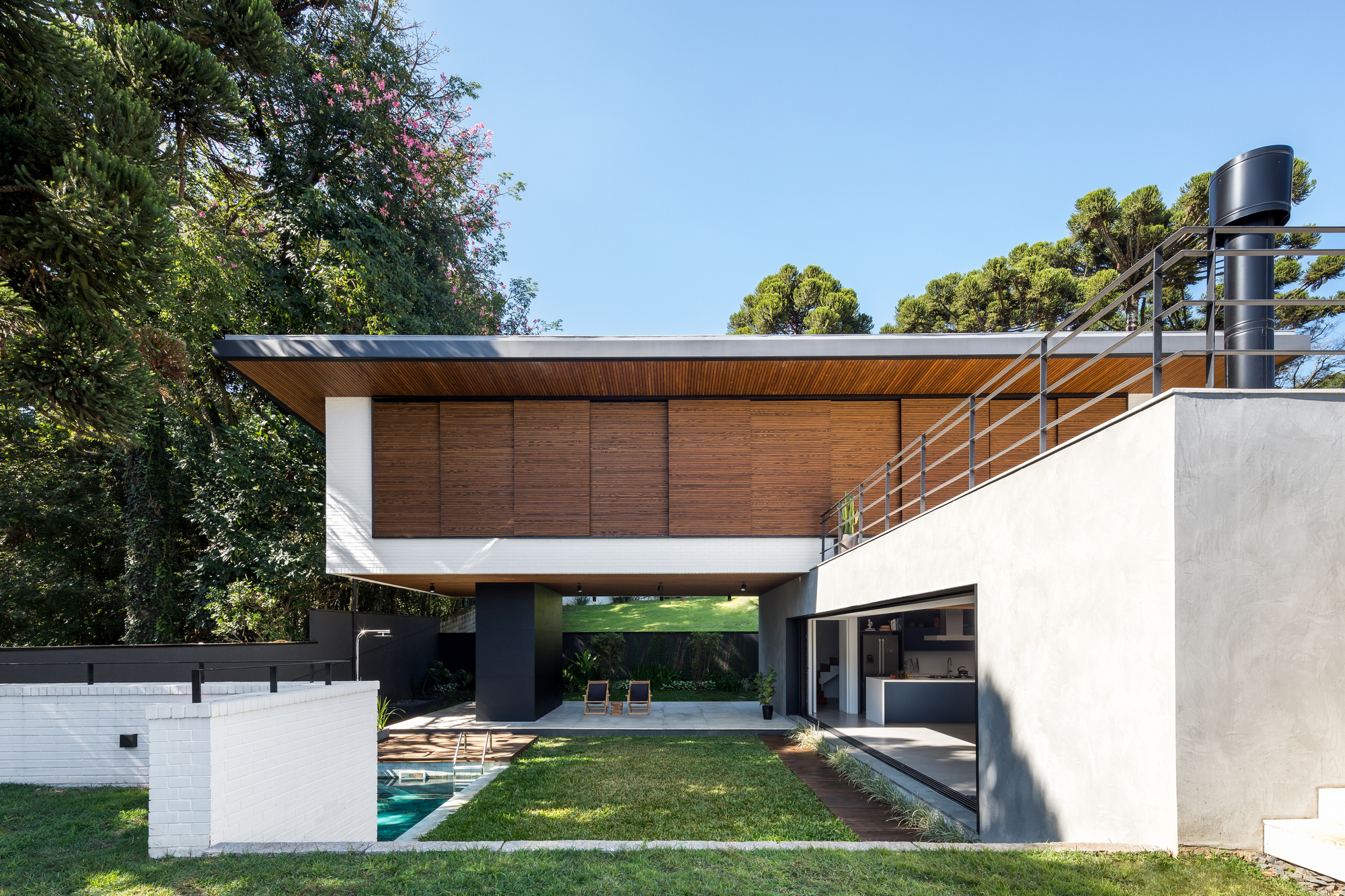 Rear elevation with a raised timber-clad volume on a dark column above a lawn and small pool, among araucaria trees