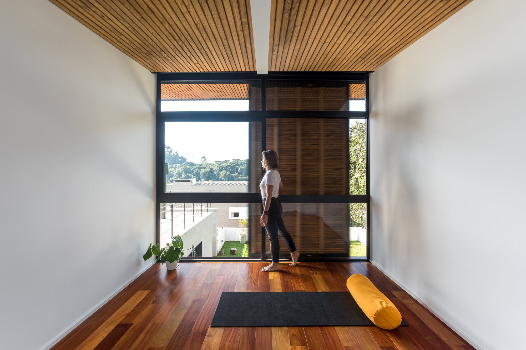 Upstairs room with a floor-to-ceiling black-framed window, slatted timber ceiling and a woman walking past a yoga mat
