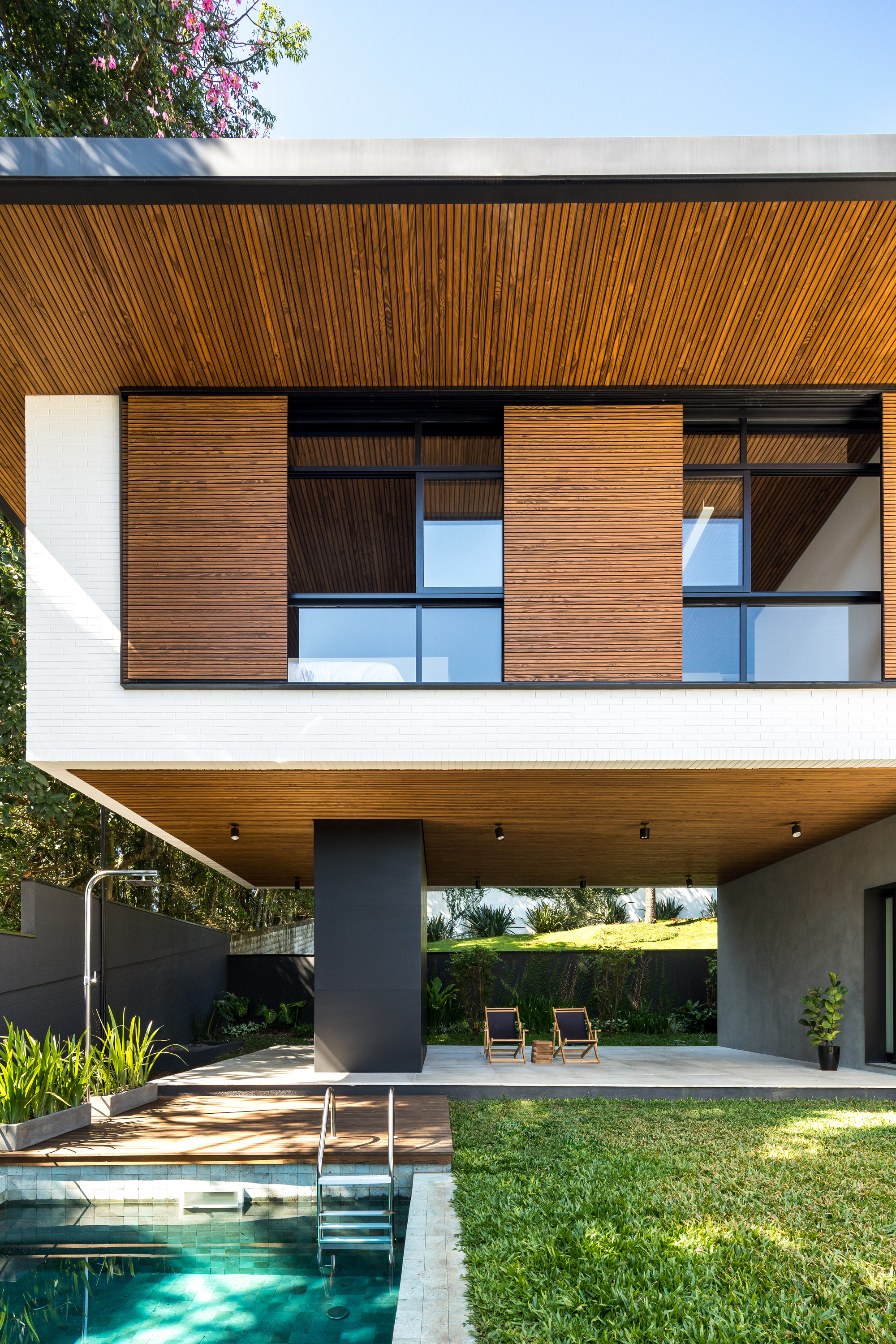 Covered terrace under the cantilevered upper floor, with timber shutters, two folding chairs and pool steps in afternoon sun