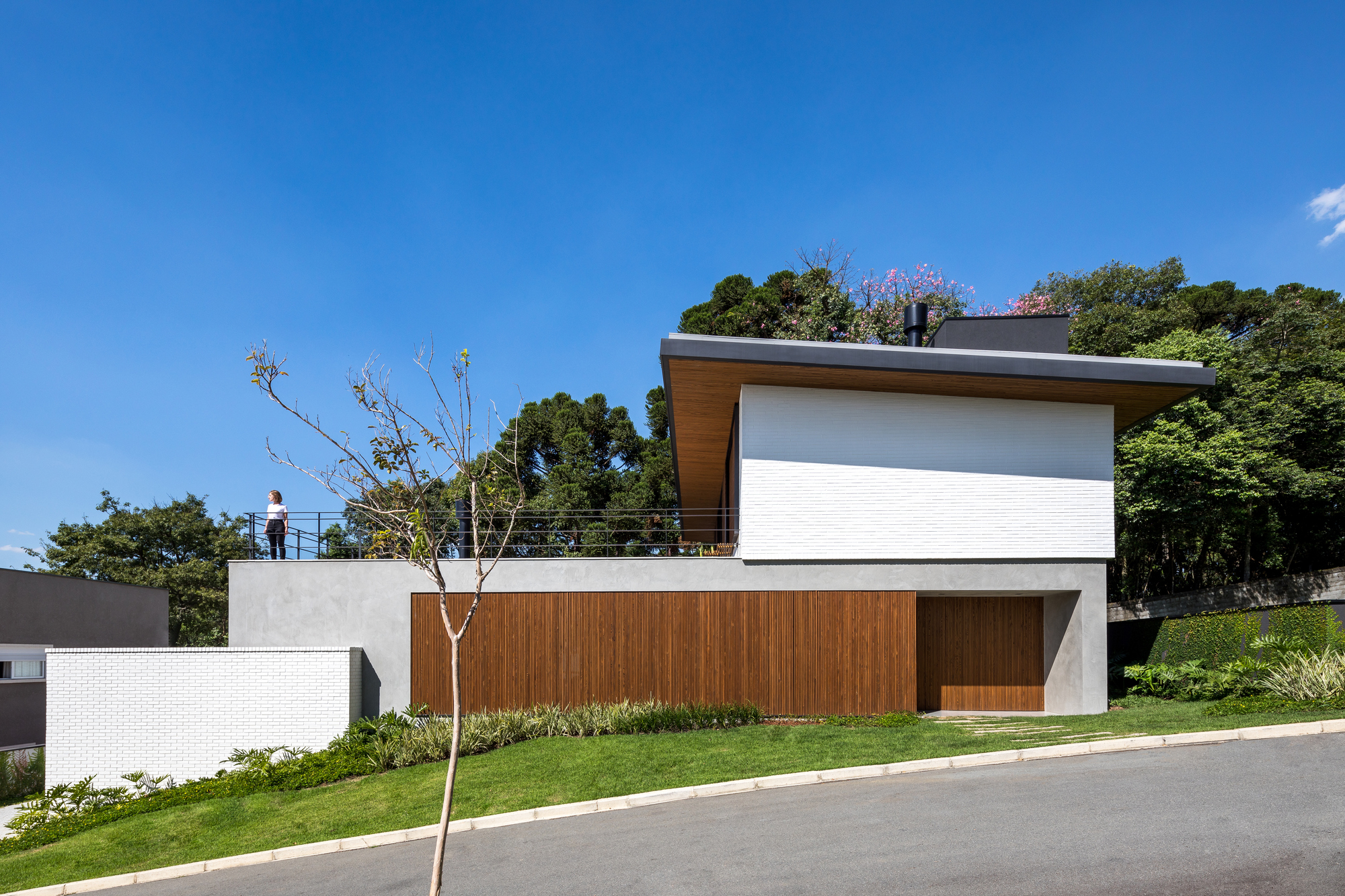 Street facade with a timber garage door below a white brick upper volume, a person standing on the roof terrace under blue sky