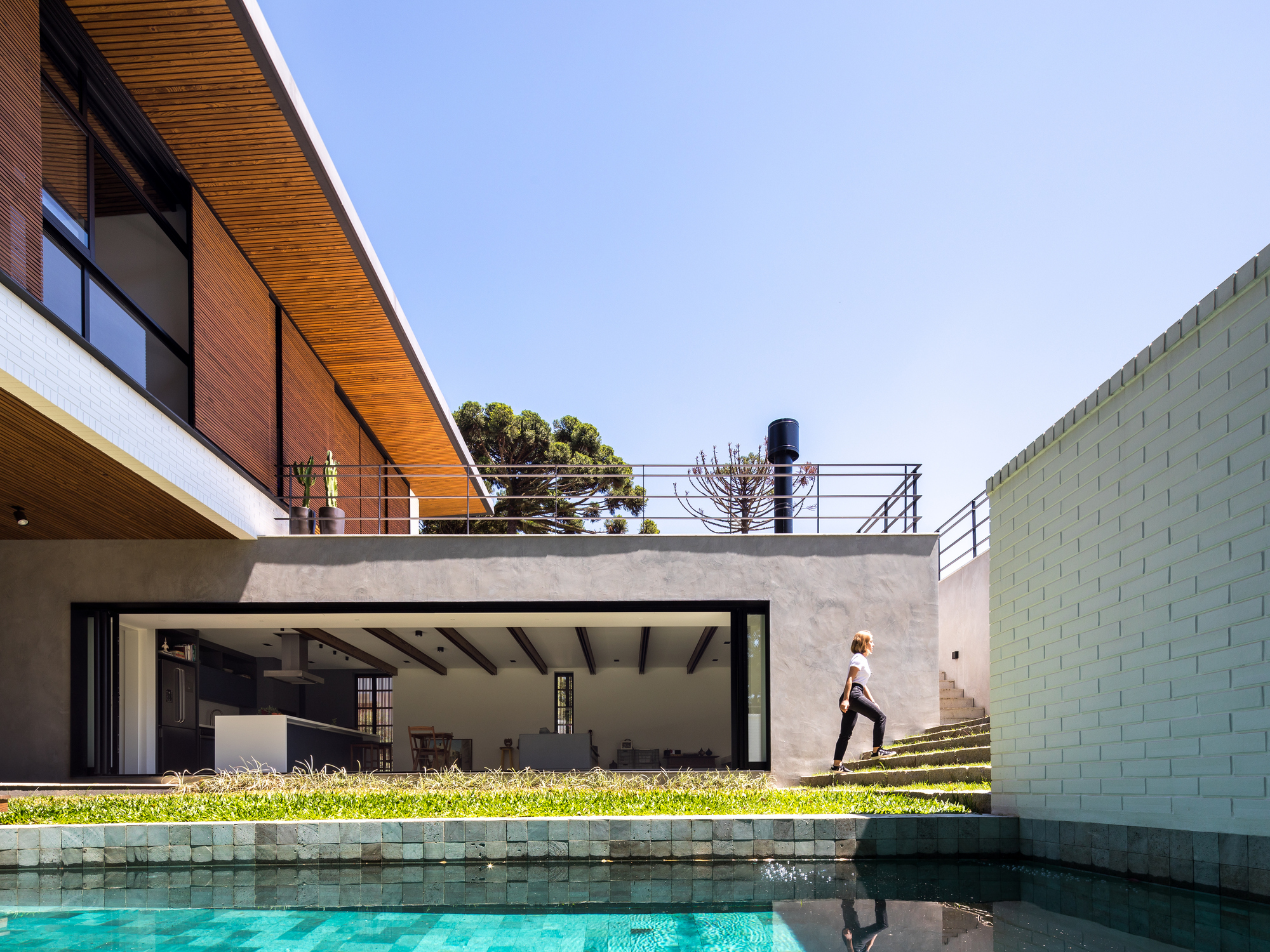 Open living room behind a grass bank and turquoise pool, with a woman climbing the grass steps in bright sun