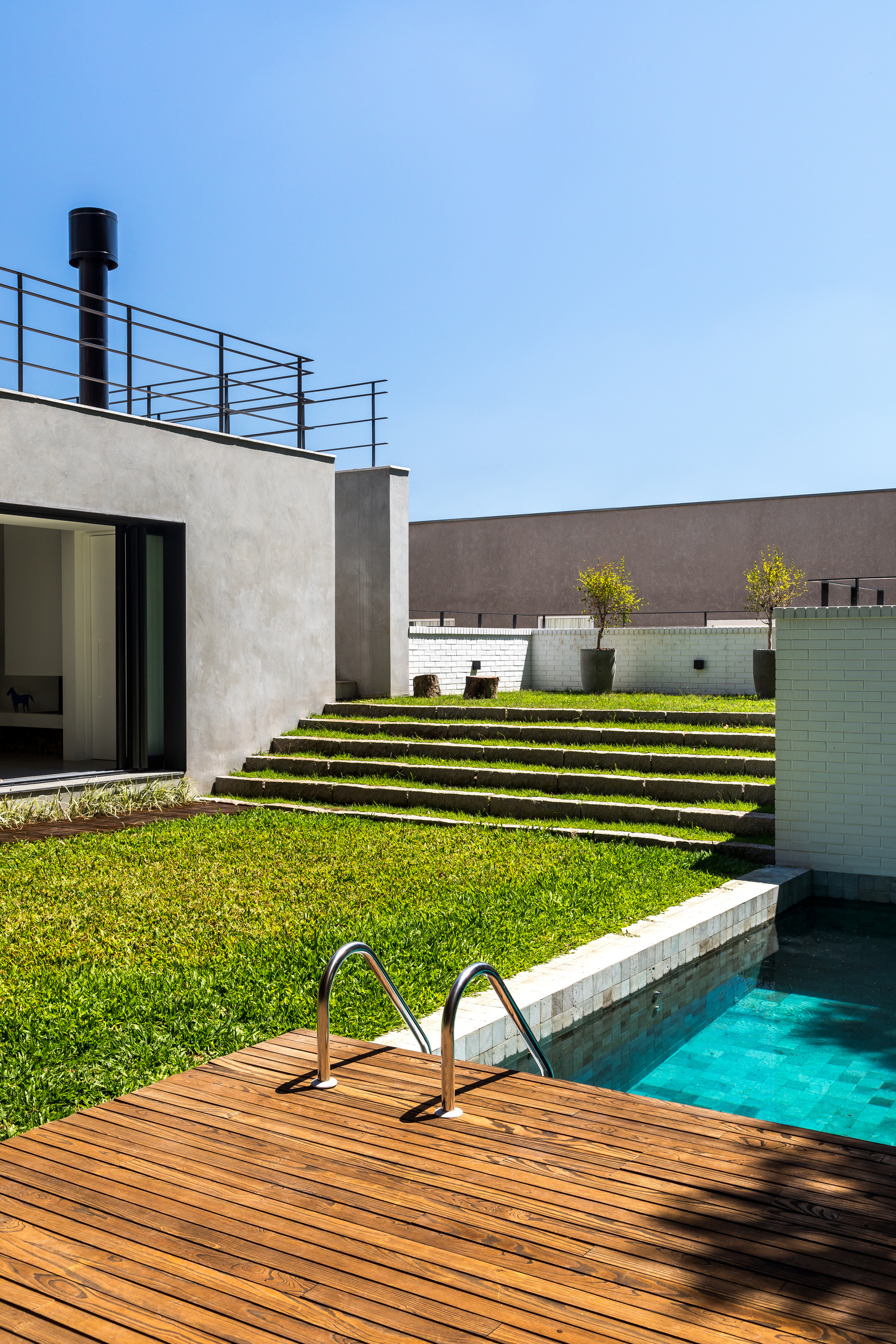 Grassed steps rising beside a turquoise pool and timber deck, with a black flue above the grey concrete wall