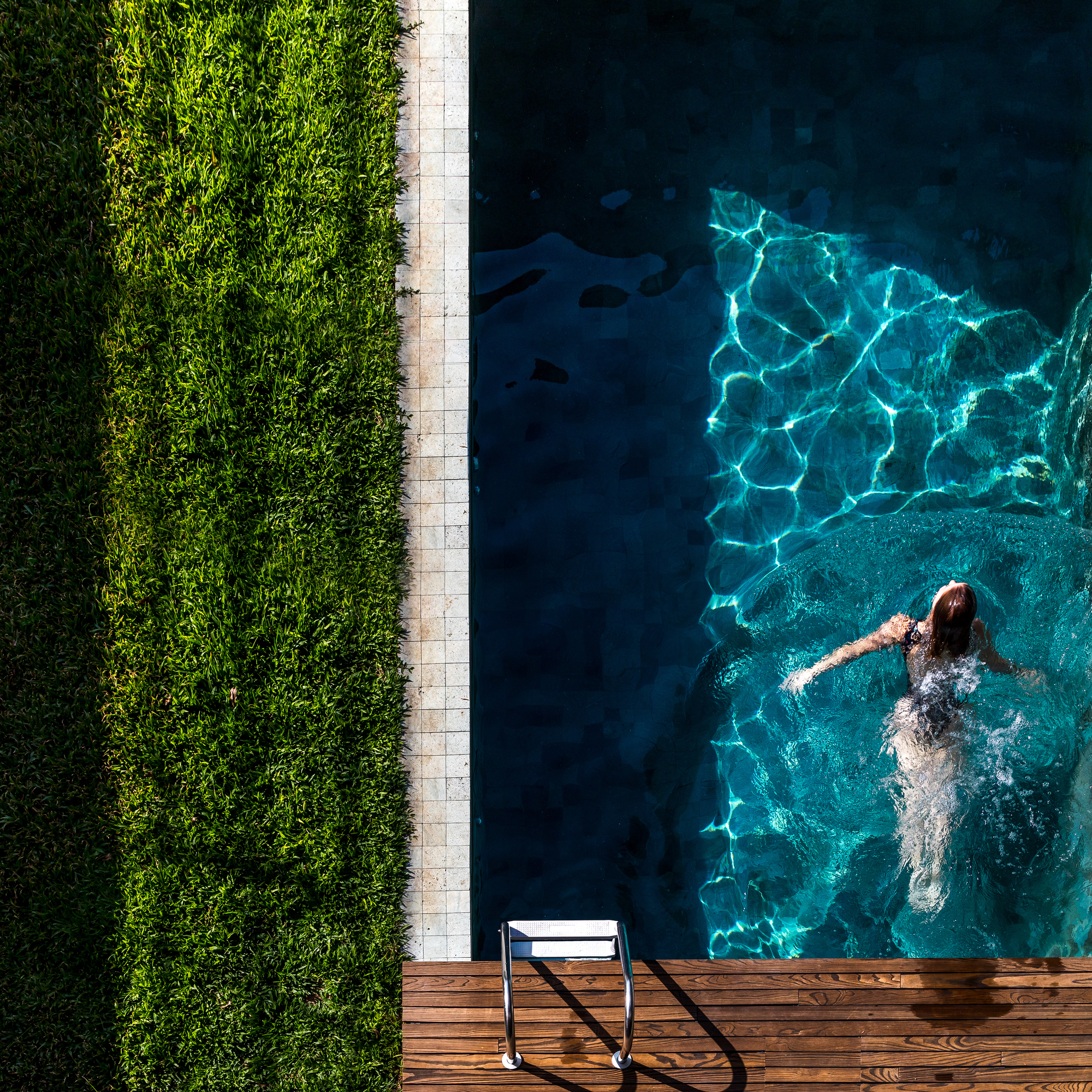 Top-down view of a swimmer in a dark pool beside a grass strip and timber deck, with light rippling on the water