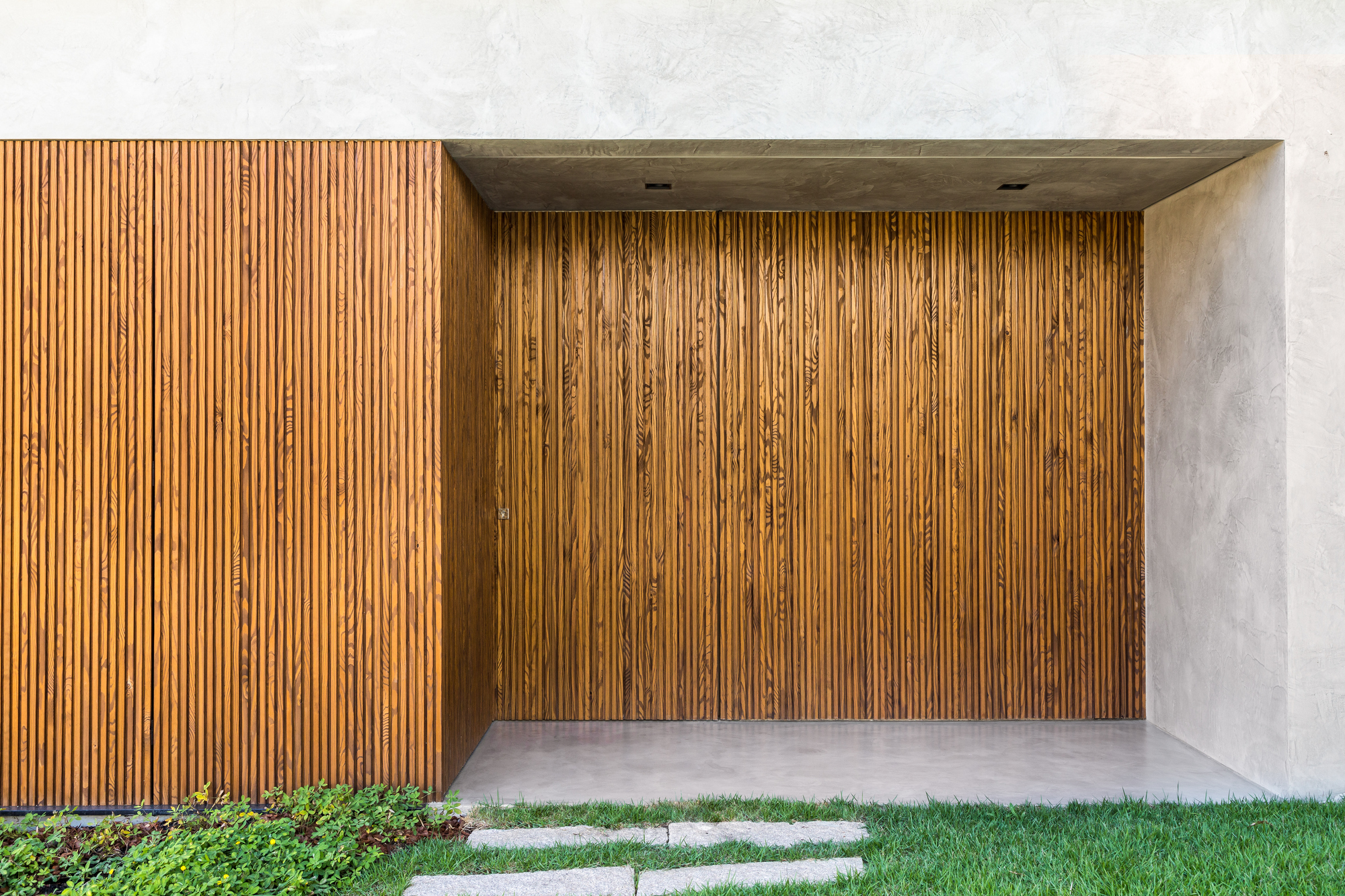 Vertical timber slat wall recessed under a concrete canopy, with stepping stones crossing the lawn in daylight