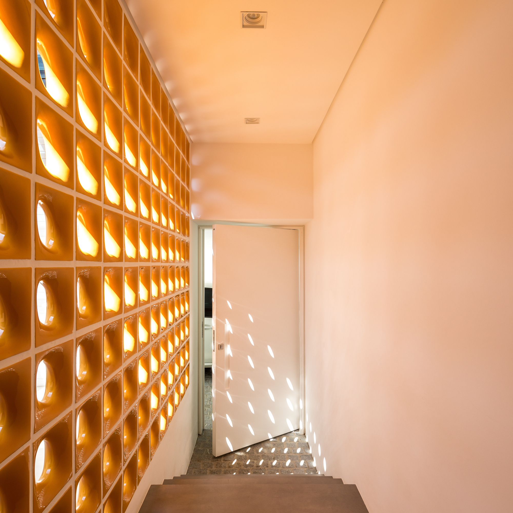 Stairwell lined with perforated ceramic block wall, throwing oval spots of sunlight across a white door and the floor
