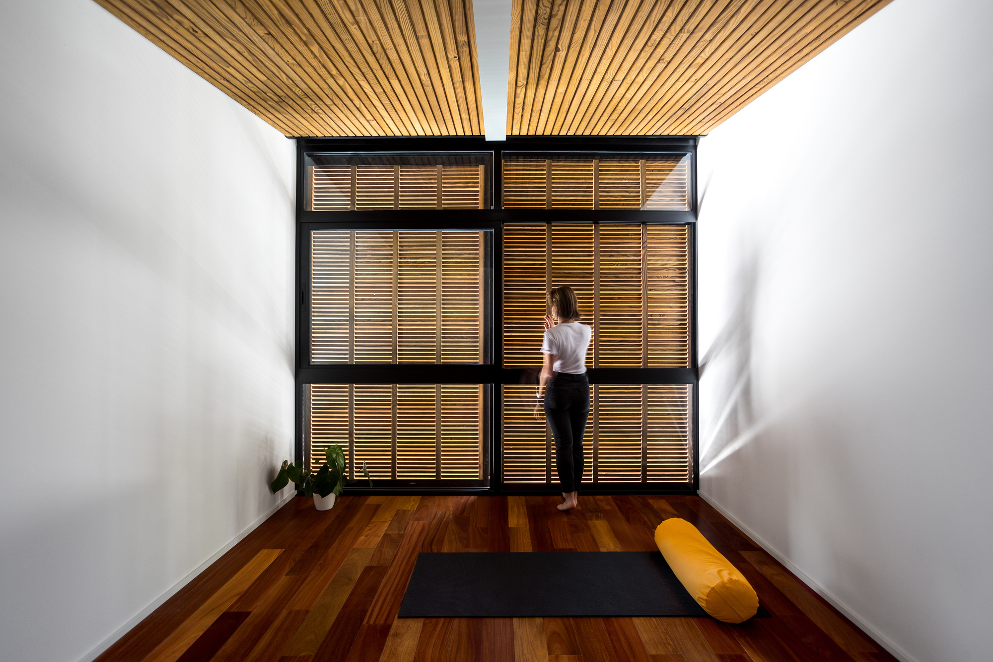 Woman standing before black-framed timber louvre shutters in a white room with a slatted ceiling and a yoga mat