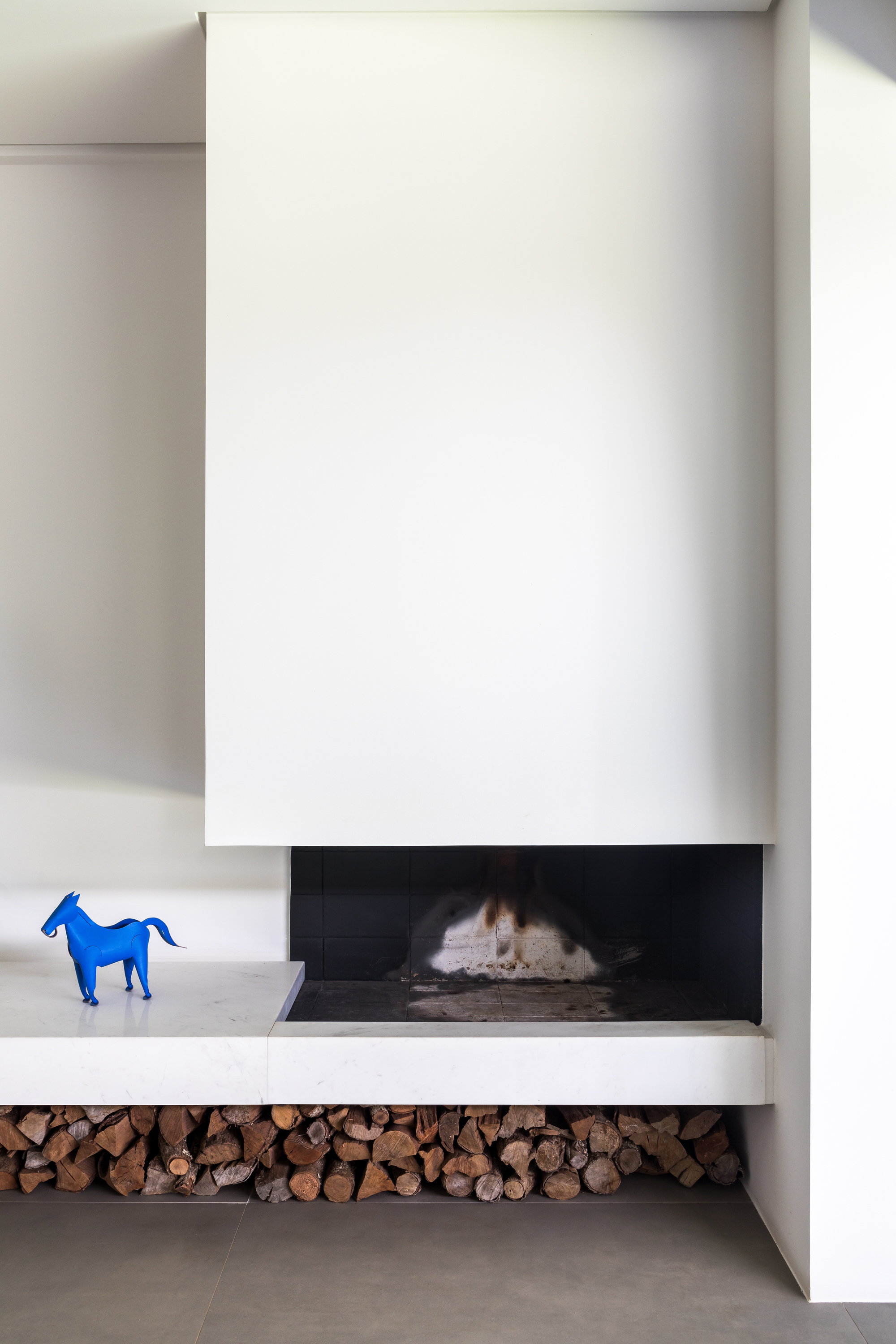 White fireplace with a cantilevered hearth ledge, stacked firewood below and a small blue horse figure on the shelf