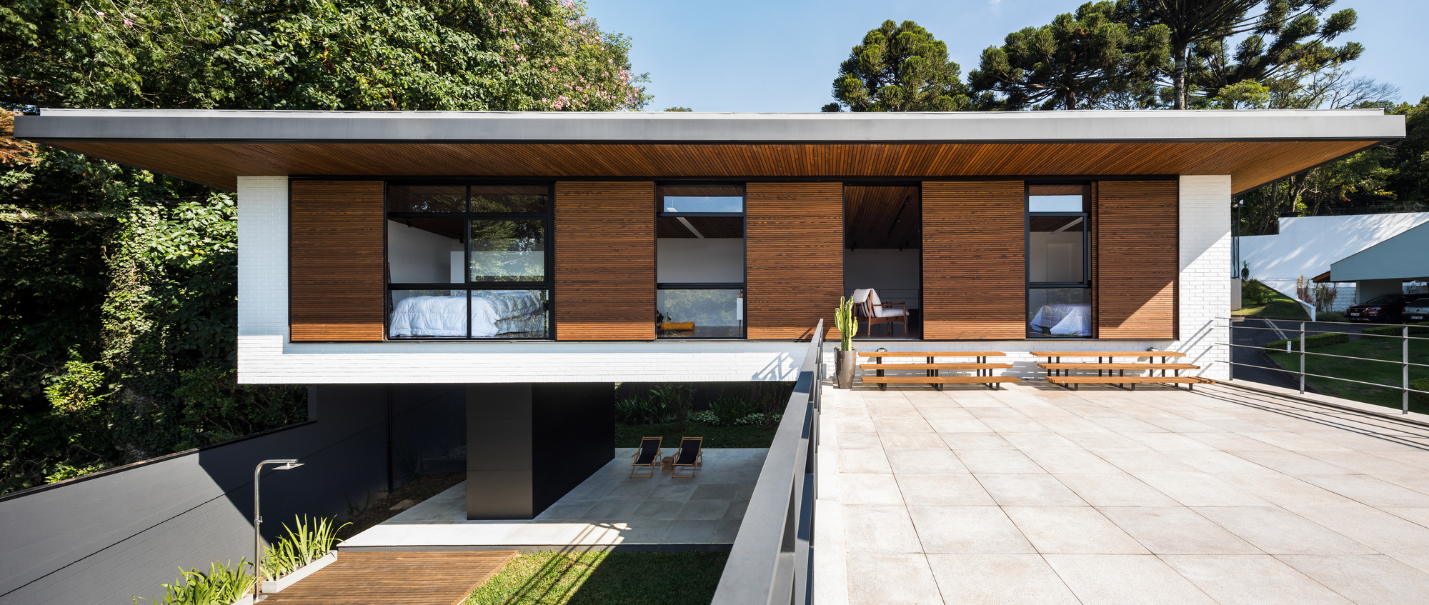 Raised upper volume with sliding timber shutters and a wood soffit, seen across a paved terrace beside tall trees