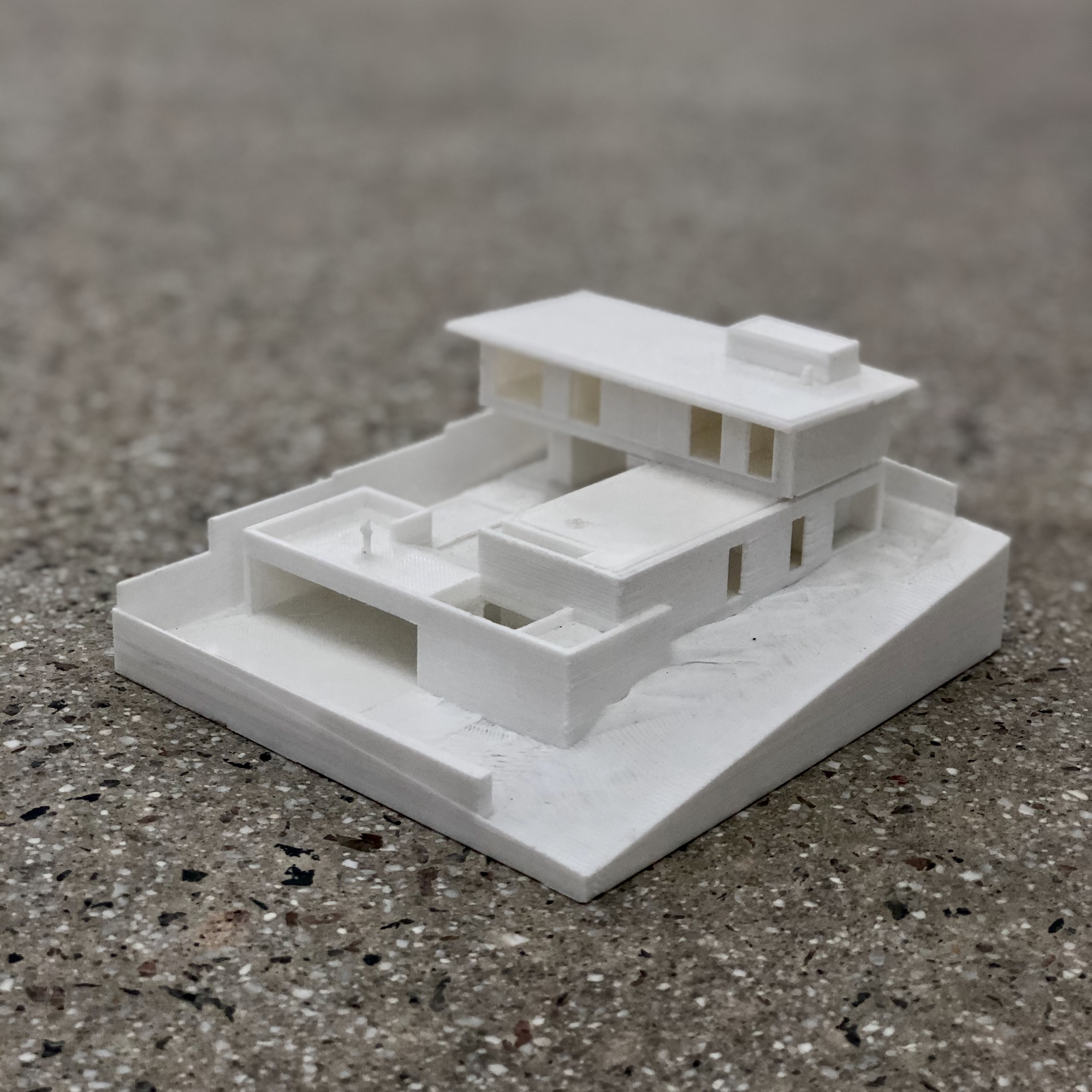 White 3D-printed site model of a two-storey house on a sloping base, resting on a speckled terrazzo floor