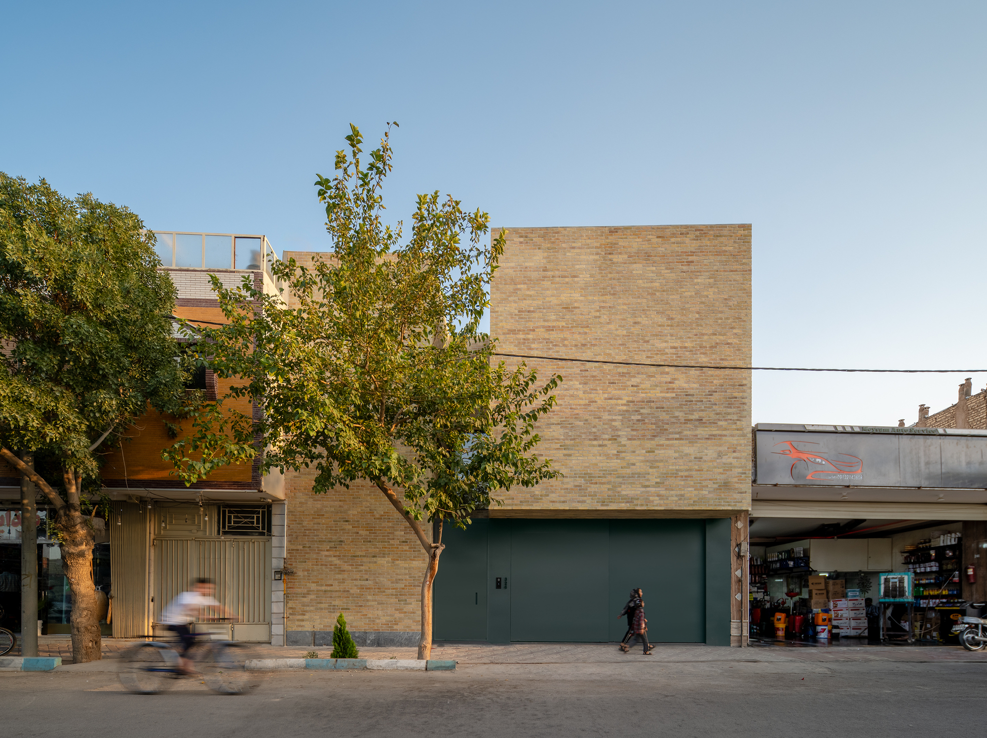 Street facade of a yellow brick block with a green steel garage door, a cyclist passing under a leafy tree