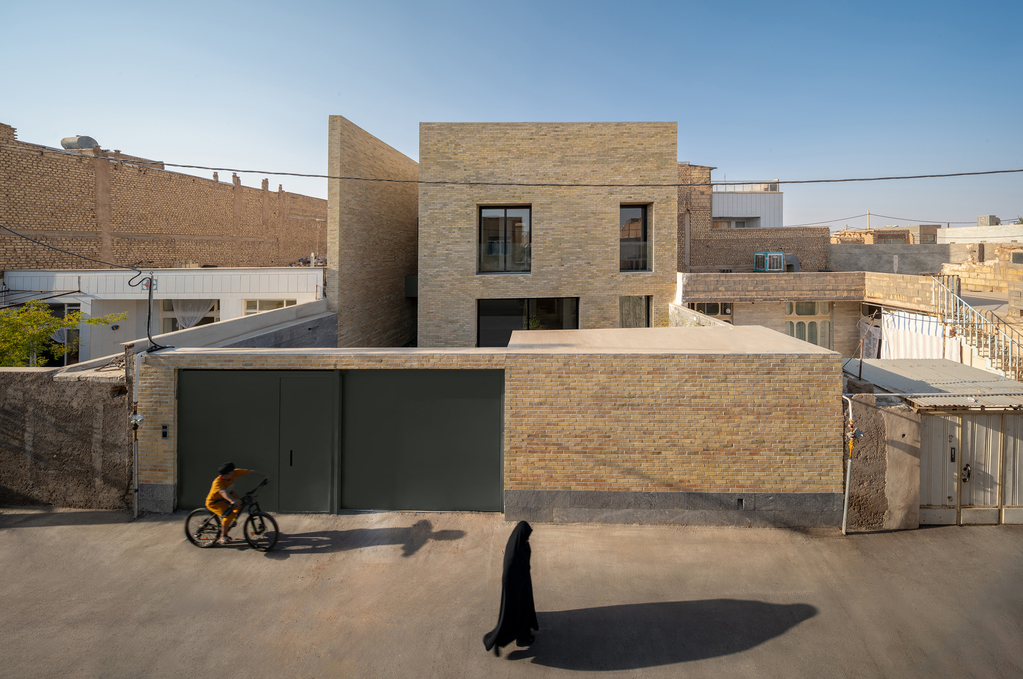 Elevated view of stepped brick volumes behind a low garden wall with green doors, a woman in black walking past