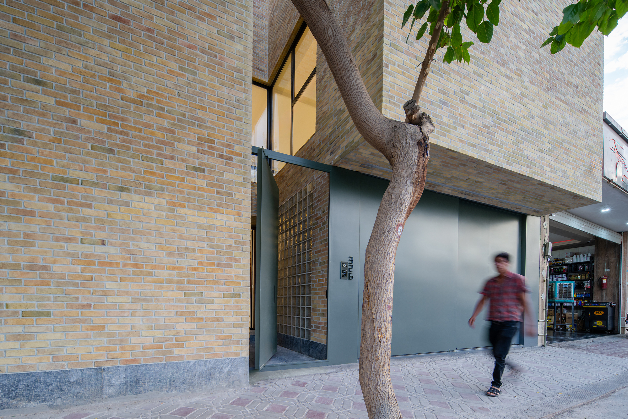 Corner entrance with a pivoting green steel door and glass block wall, a blurred pedestrian passing a leaning tree trunk