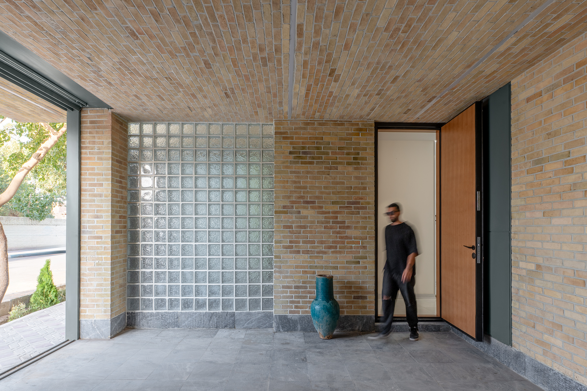 Covered entry porch with a brick vaulted ceiling, glass block panel, teal ceramic vase and open timber door in afternoon light
