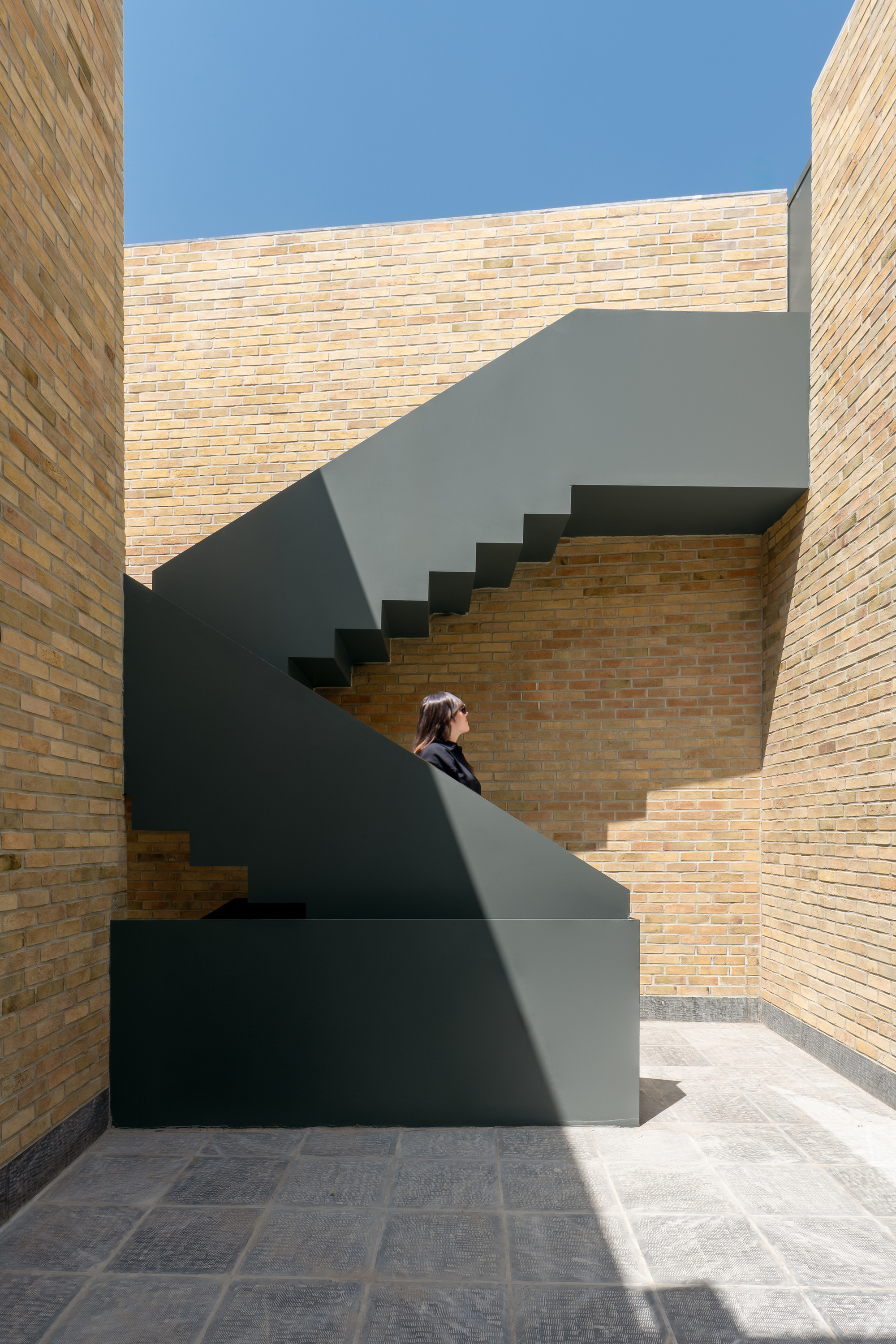 Green steel staircase rising between tall brick walls in a courtyard, a woman in shadow under a clear blue sky