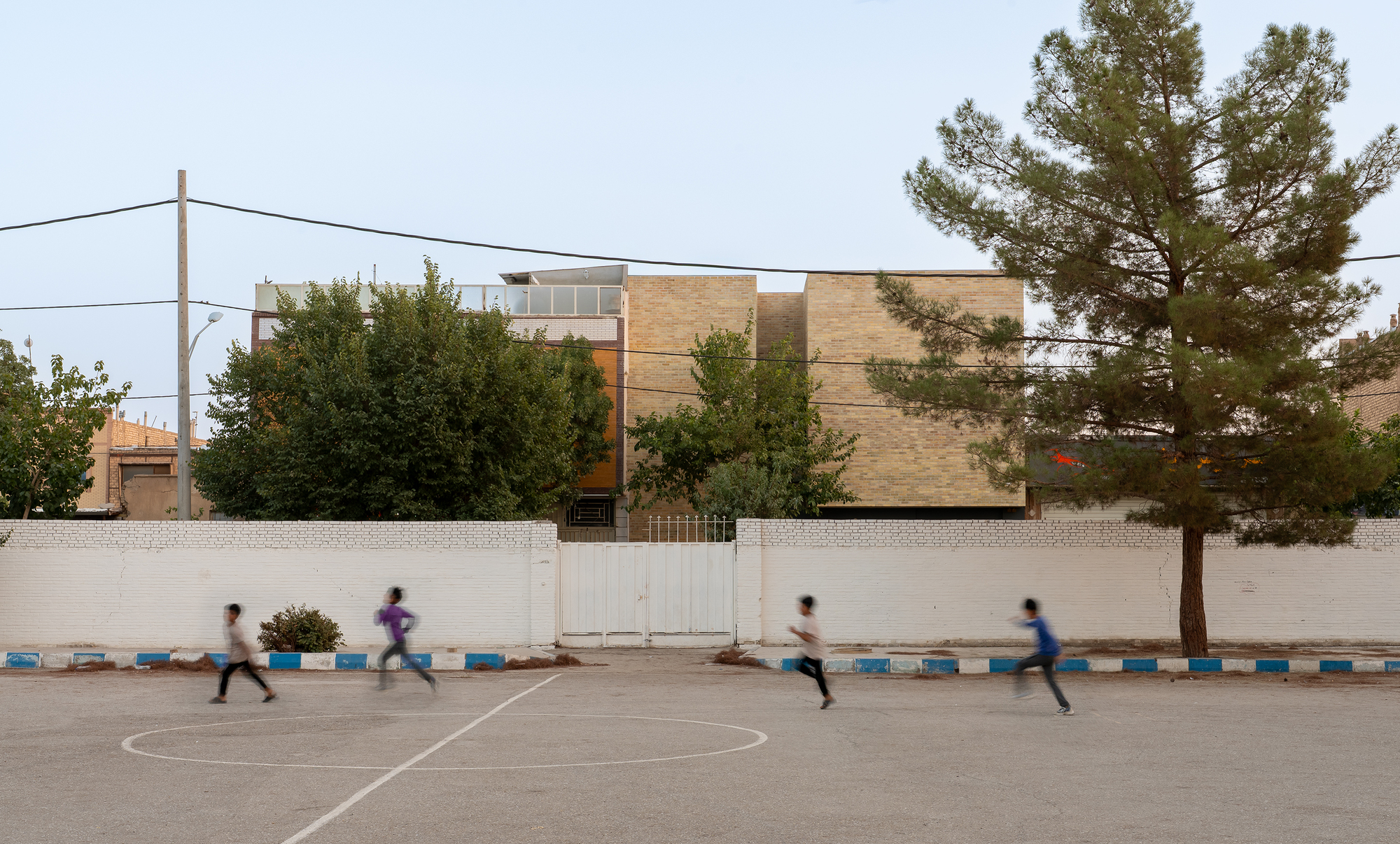 Brick house seen over a white perimeter wall from a paved court, children running past a pine tree at dusk