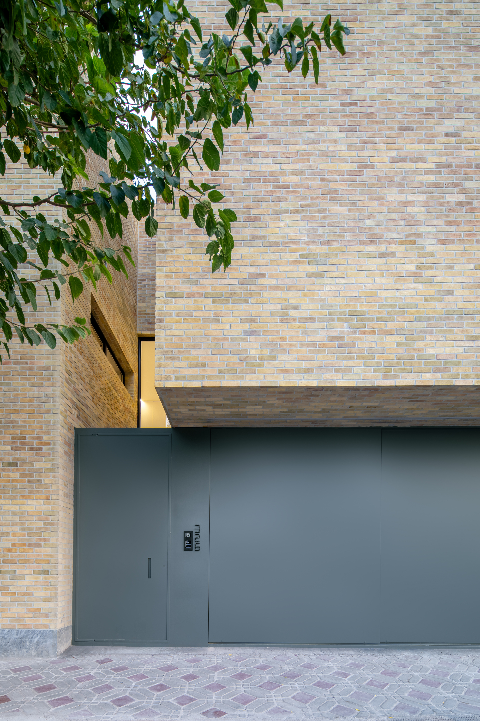 Grey steel garage door and pedestrian gate beneath a cantilevered brick volume, with leafy branches hanging across the corner