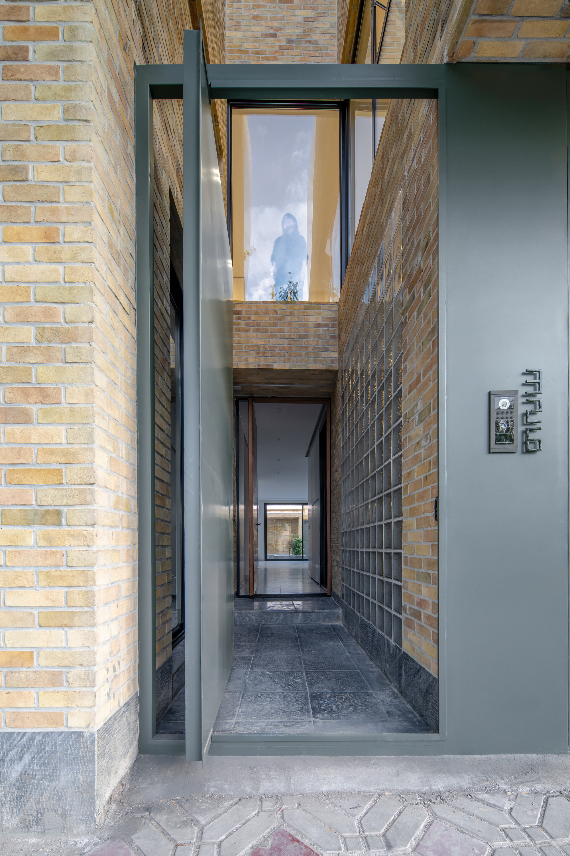 Open pivot entry door framing a narrow slate-paved passage between brick and glass block walls, with a figure visible in the window above