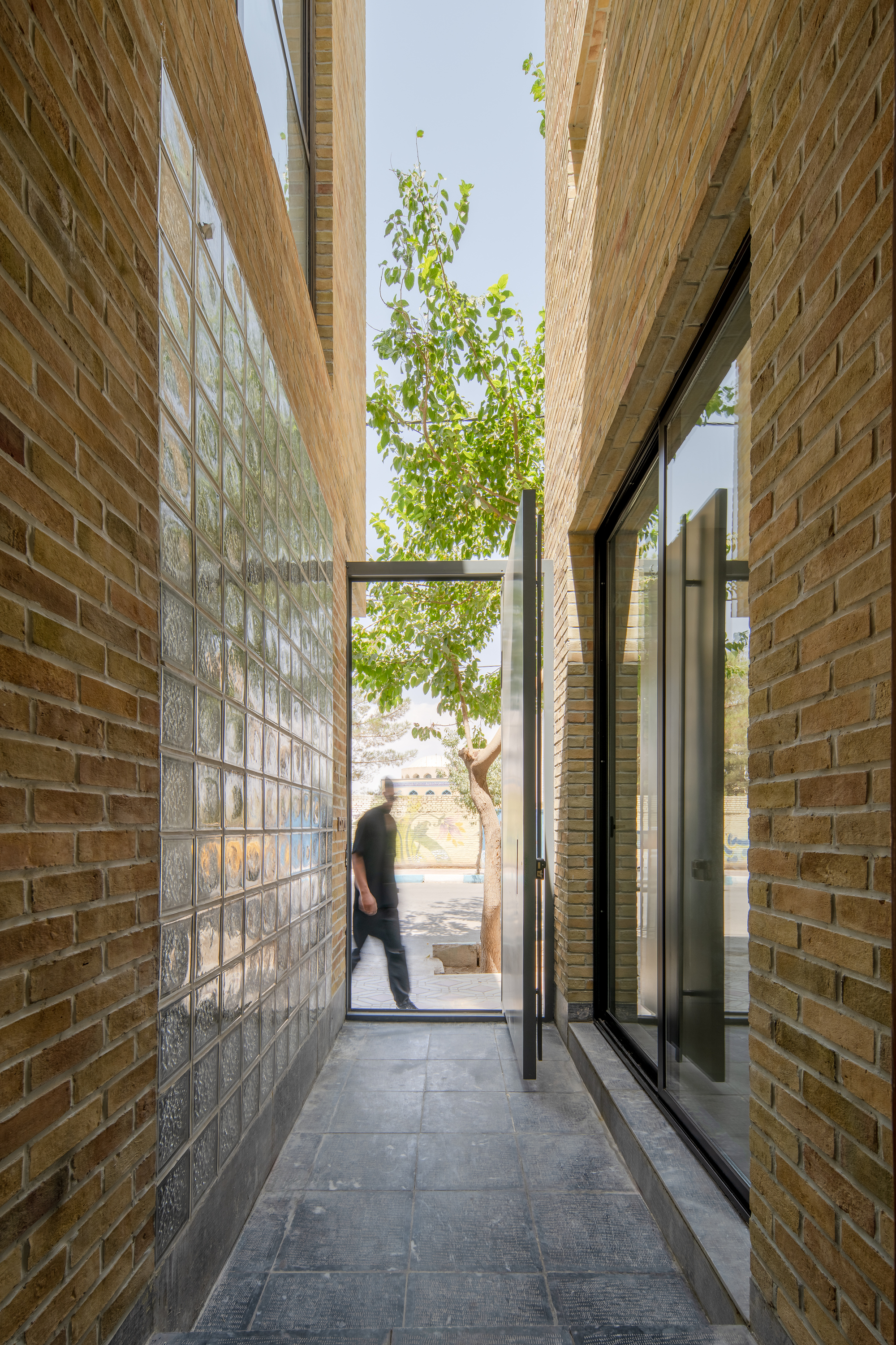 Slate-floored brick passage lined with glass block, opening to a sunlit street where a man walks past under a tree