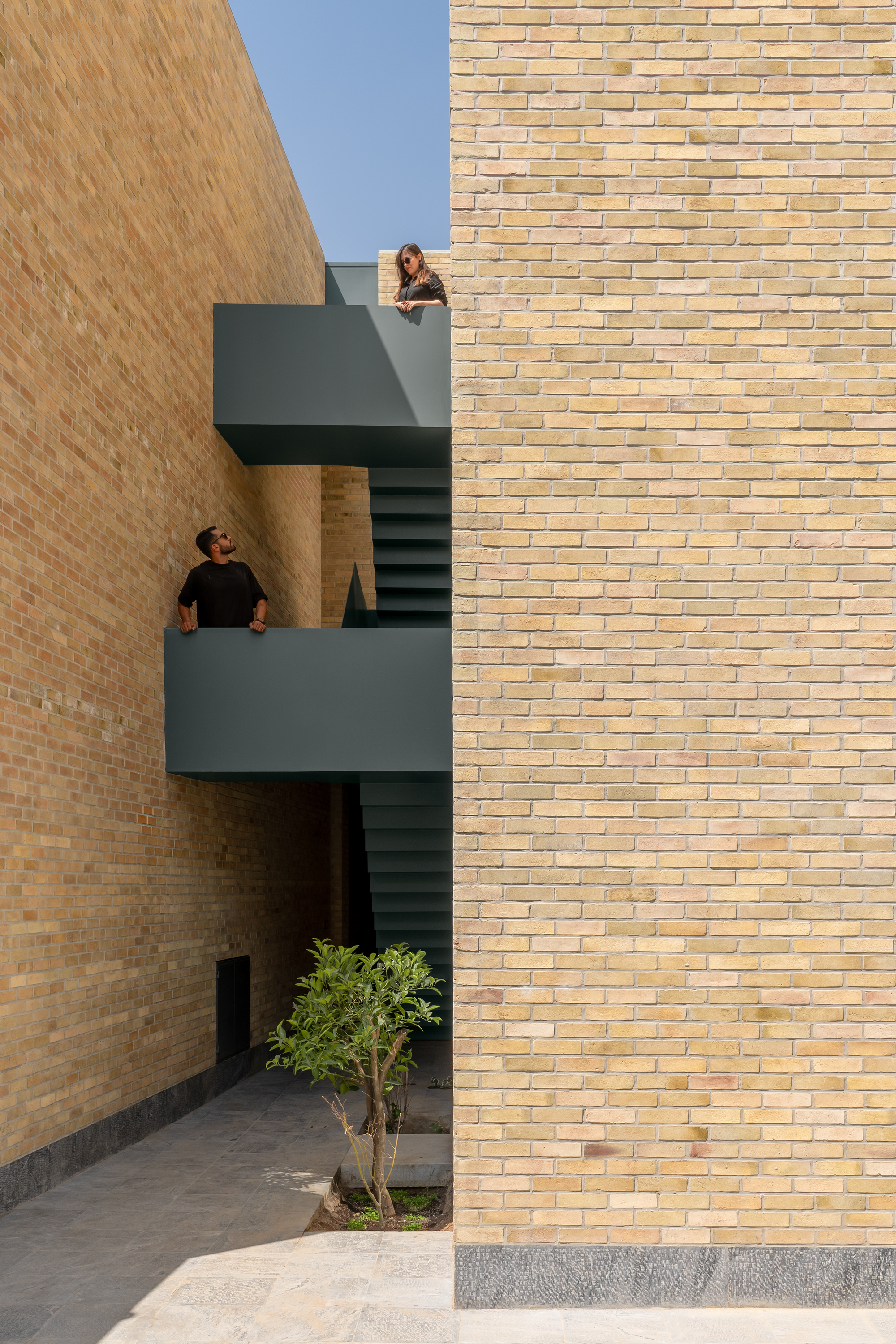 Grey steel stair landings between two brick walls, with two people leaning on the rails under a clear blue sky