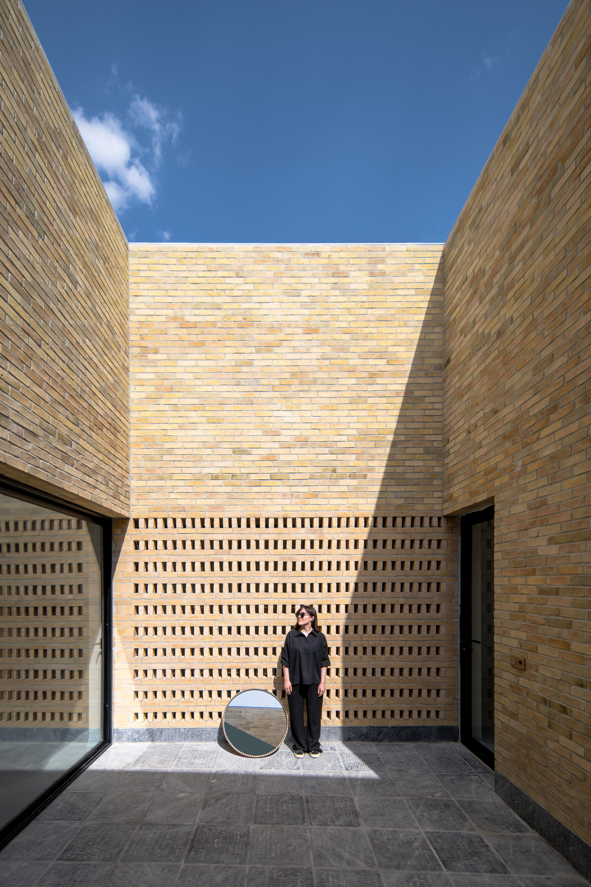 Brick courtyard with a perforated lattice wall, a woman standing beside a round mirror in strong sunlight and hard shadow