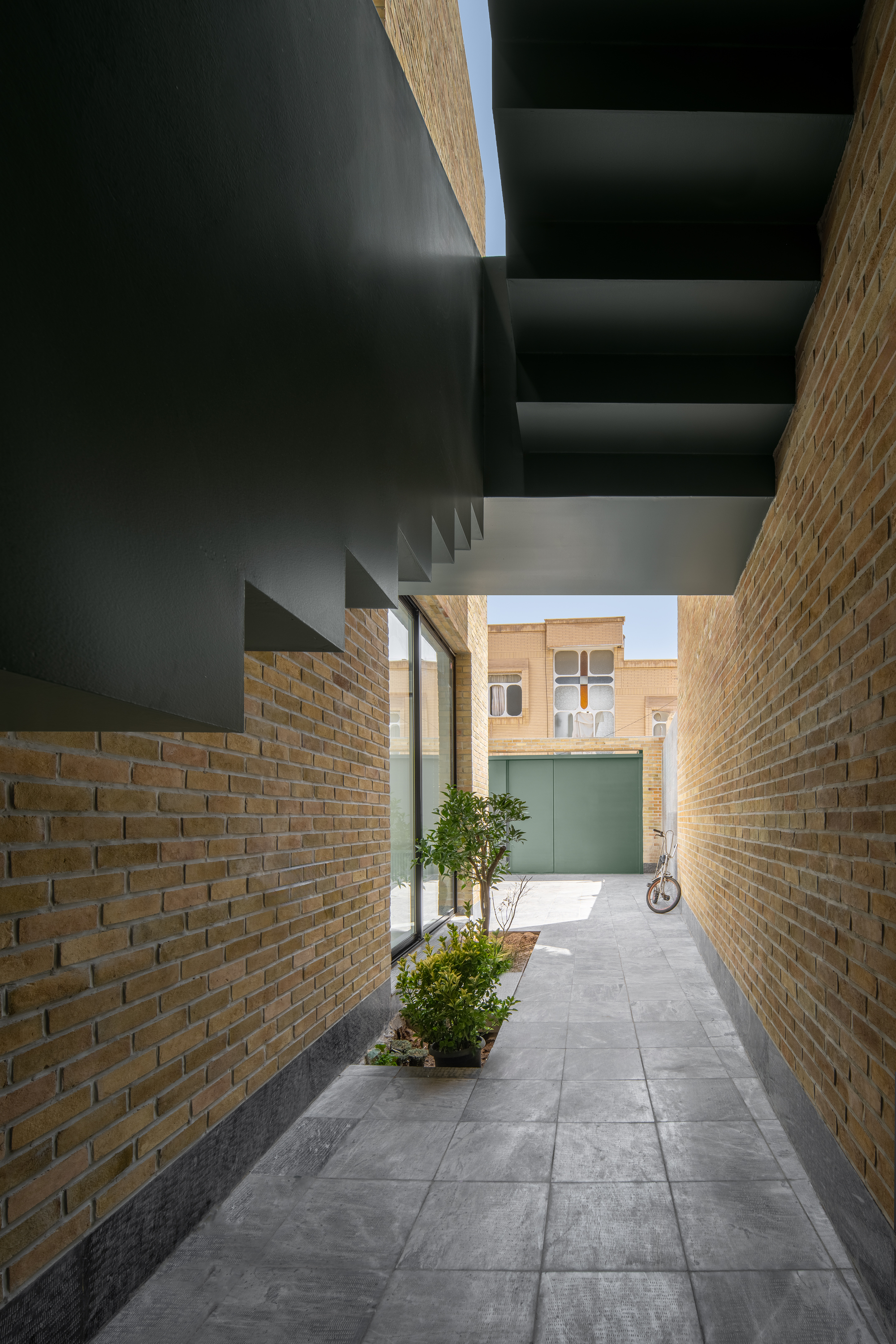 Dark steel stair soffit stepping over a narrow brick alley with small planted bed and a bicycle leaning near the far green gate