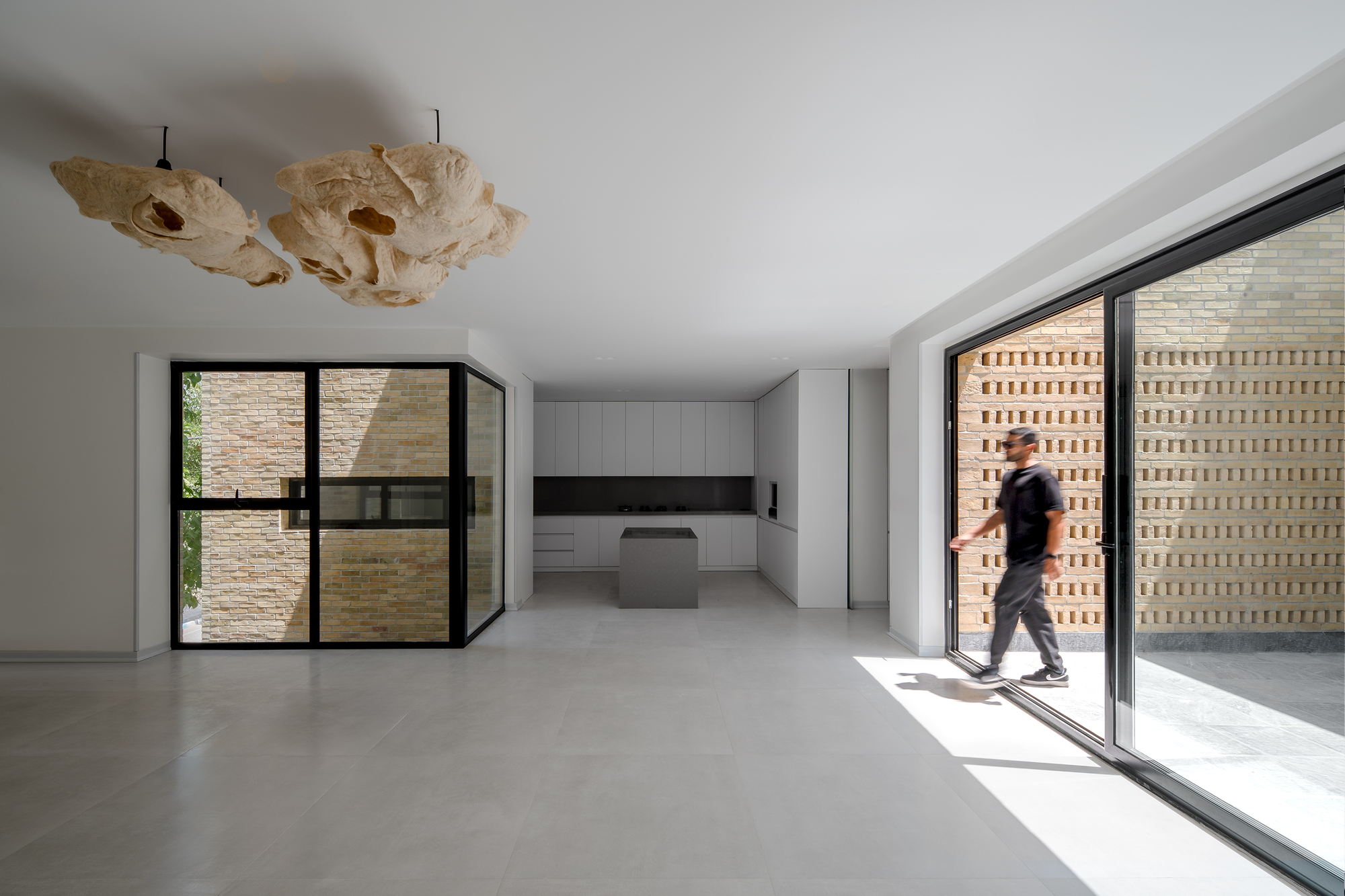Open living area with sculptural pendant lights and a sliding glass door onto a perforated brick wall, a man stepping through in sunlight