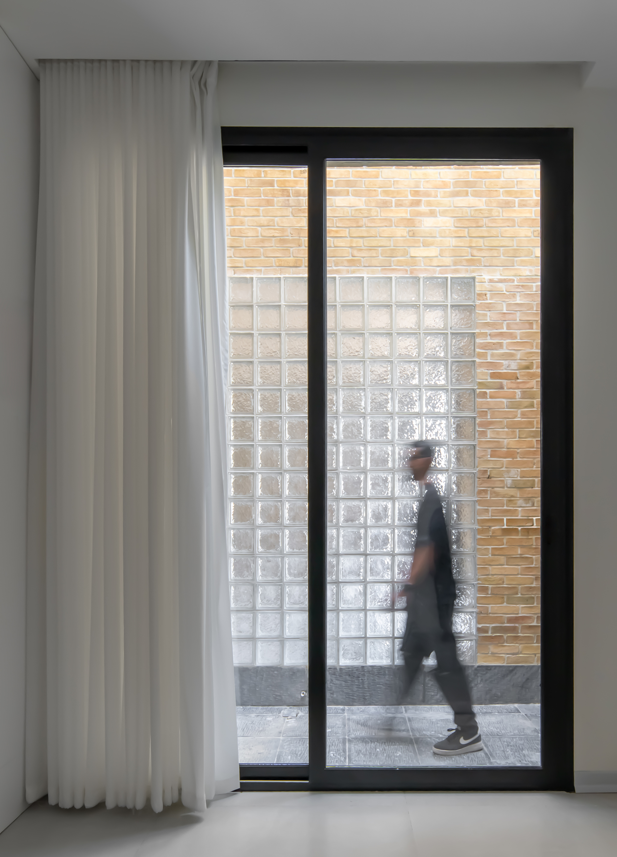 Sliding glass door framing a glass block panel set into a brick wall, a blurred walking figure and sheer curtain at left