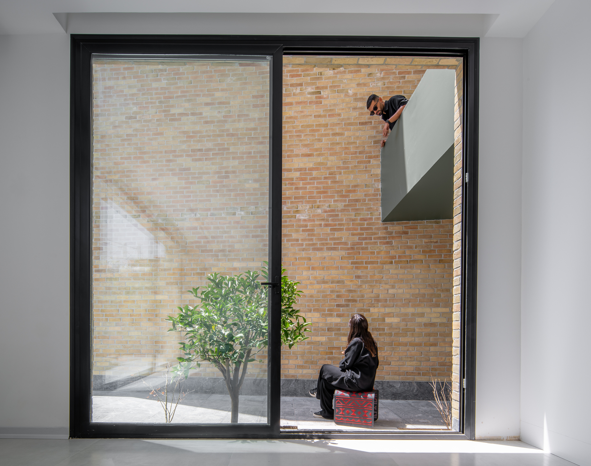 Brick courtyard seen through a black-framed glass door, with a potted citrus tree, a seated woman and a man leaning from a green balcony