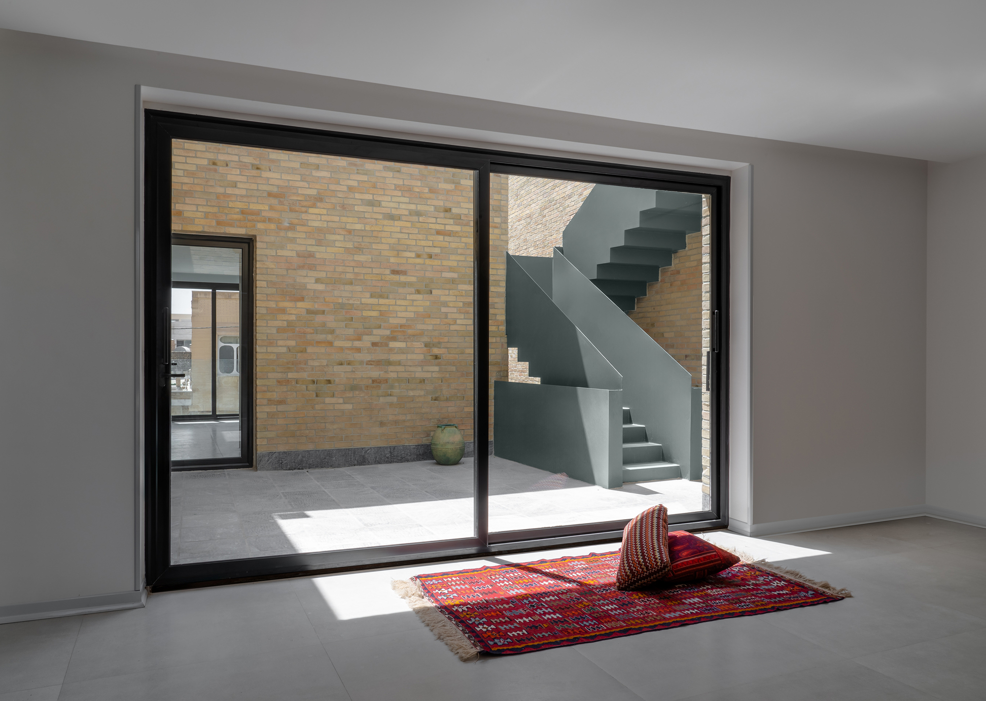 Sliding glass door opening to a brick courtyard with a green concrete stair, a red kilim rug and cushions in a patch of sun
