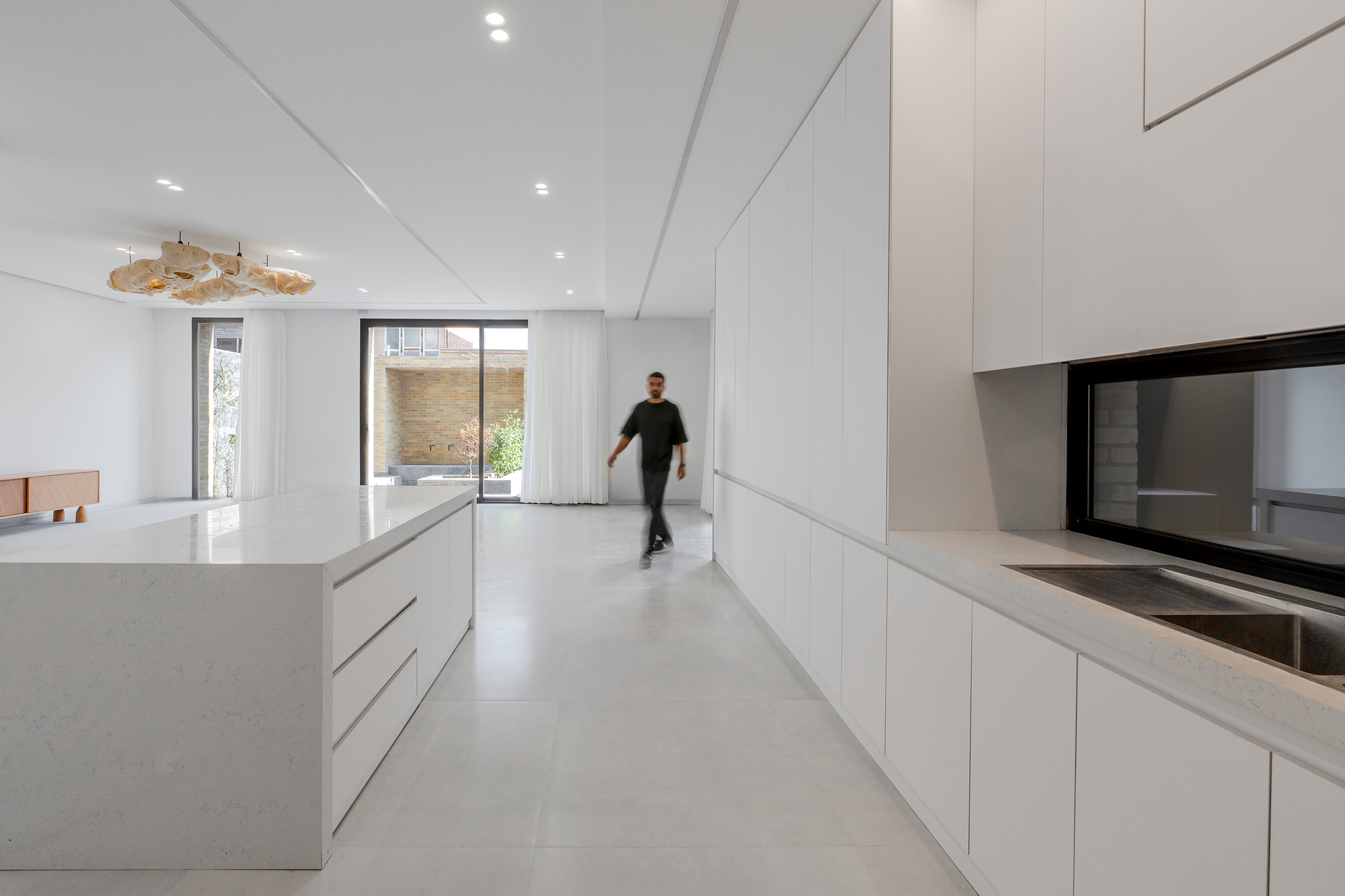 Long kitchen with white cabinetry, a stone island and a pass-through window, a man walking toward curtained glass doors in daylight