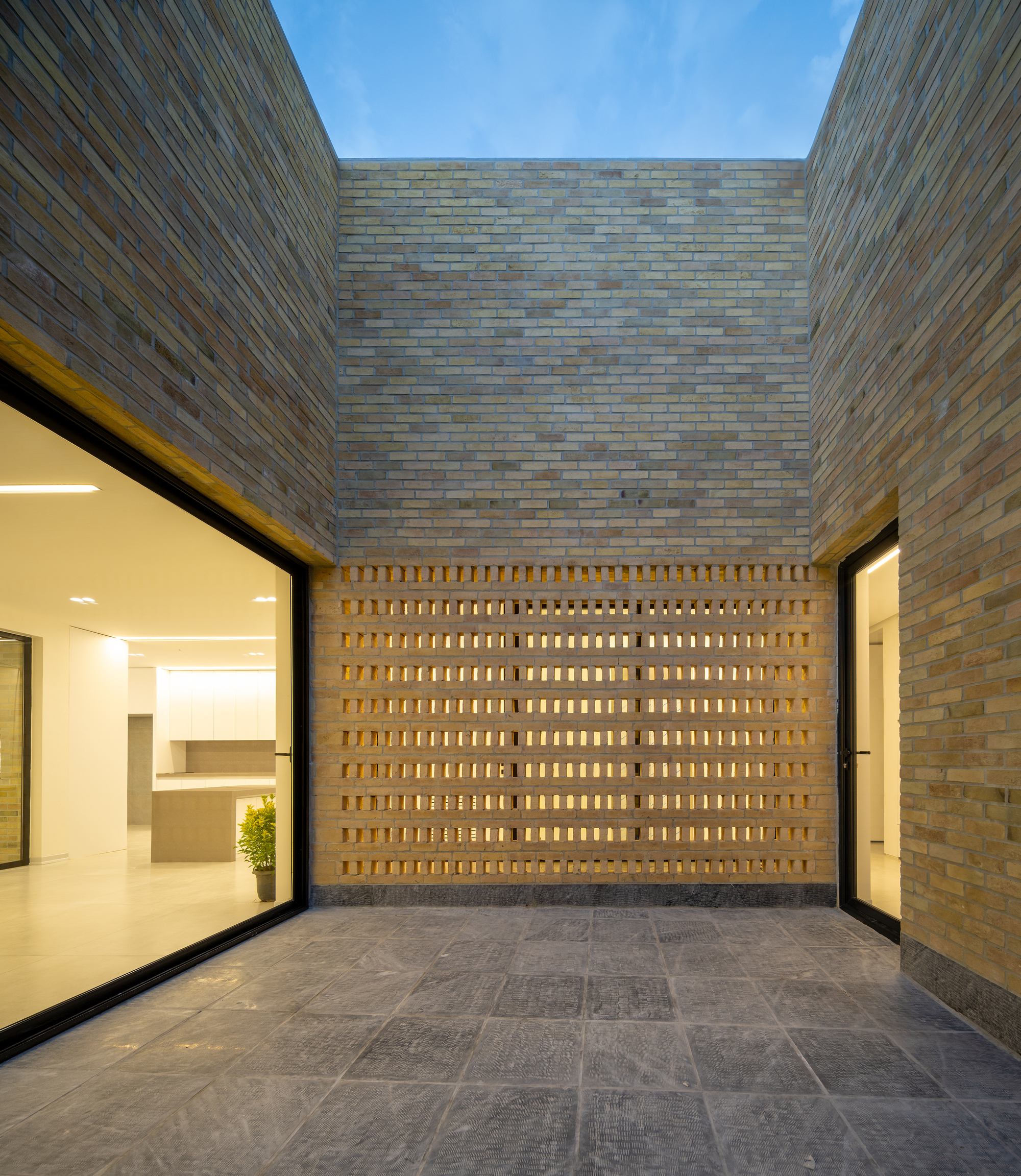 Brick courtyard with a perforated screen wall glowing from behind, framed by glazed openings and grey stone paving at dusk