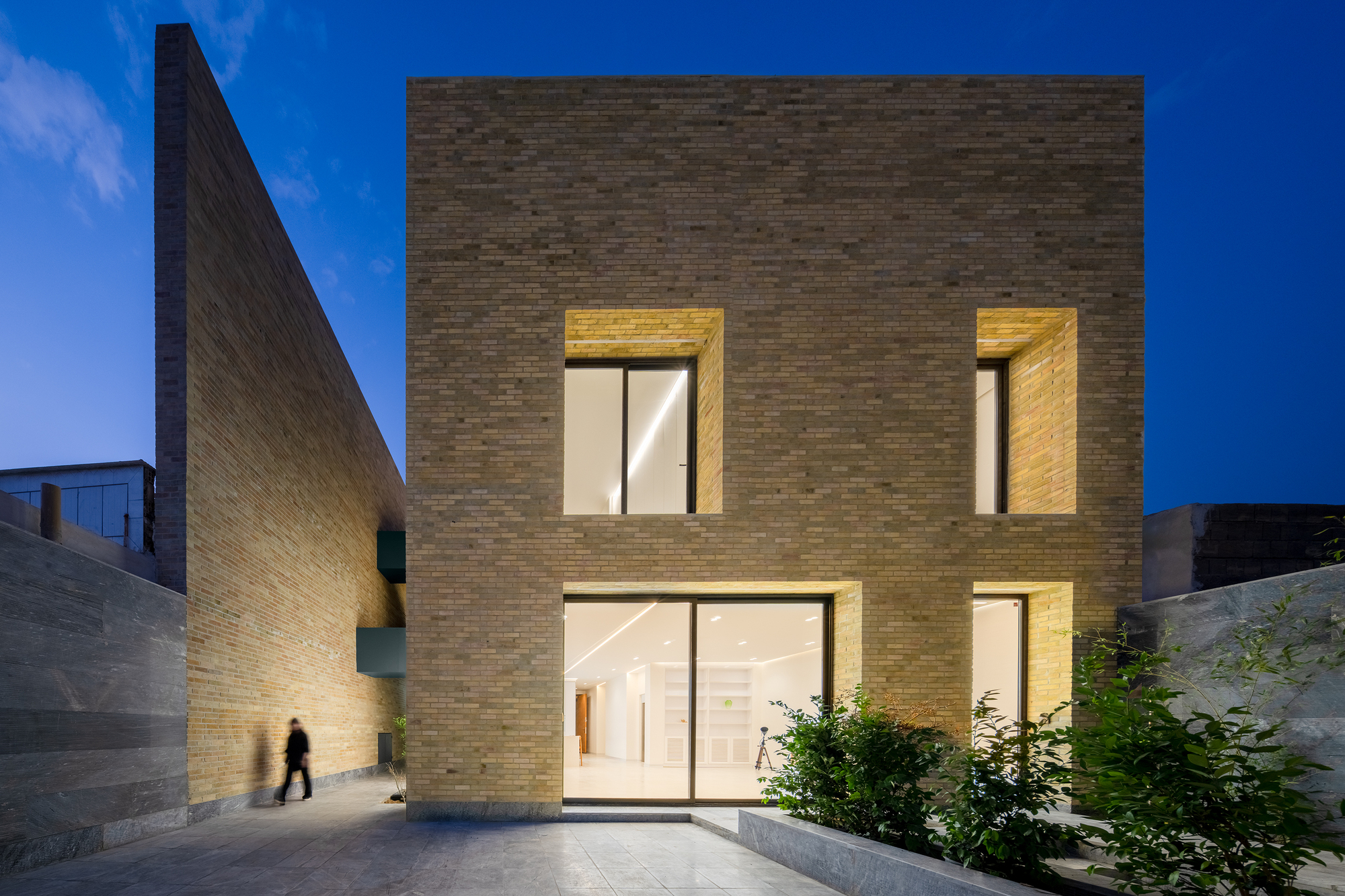 Brick house facade with deep recessed windows lit from within, a pedestrian passing the angled side wall at blue hour