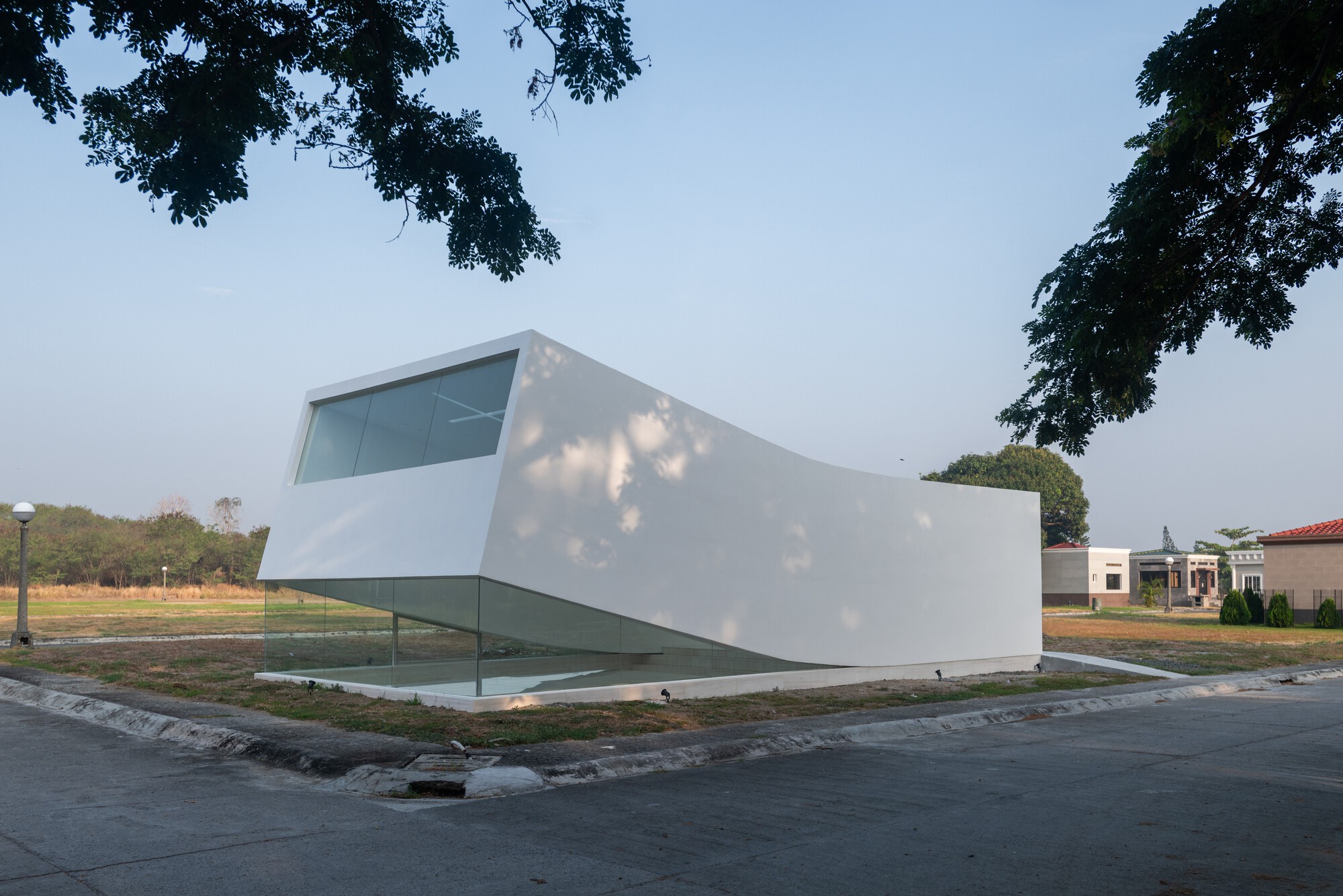 Corner view of the white tilted volume with a glazed base, dappled by tree shadows on a clear afternoon