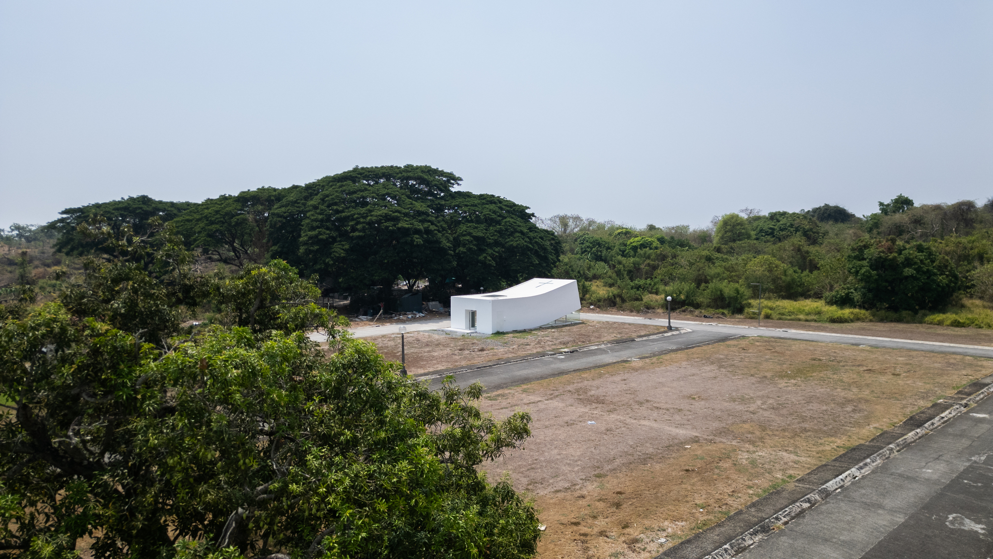 Elevated view of the small white building beside a large rain tree and dry grass lot in hazy light