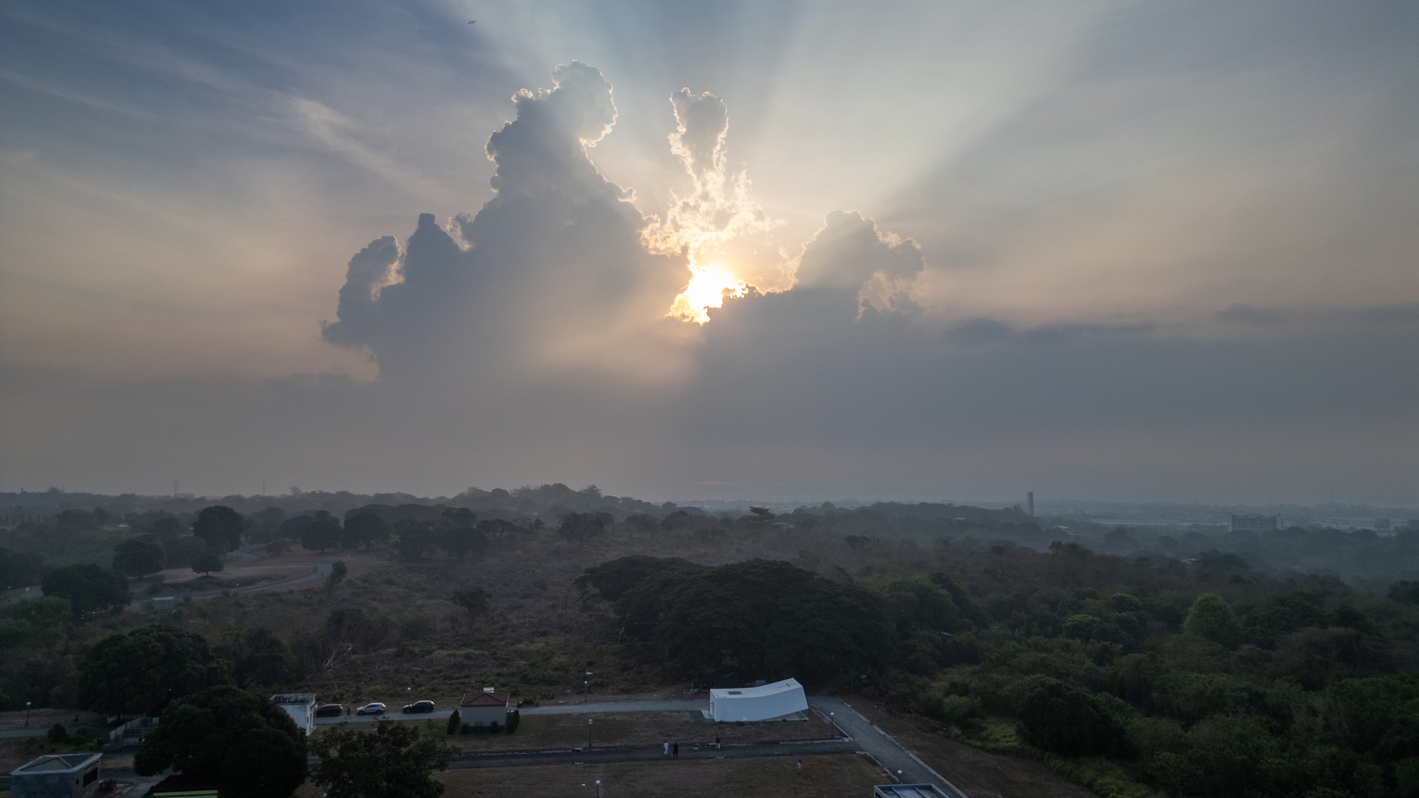 Aerial view of the white building among trees below a sunburst breaking through heavy clouds at dusk