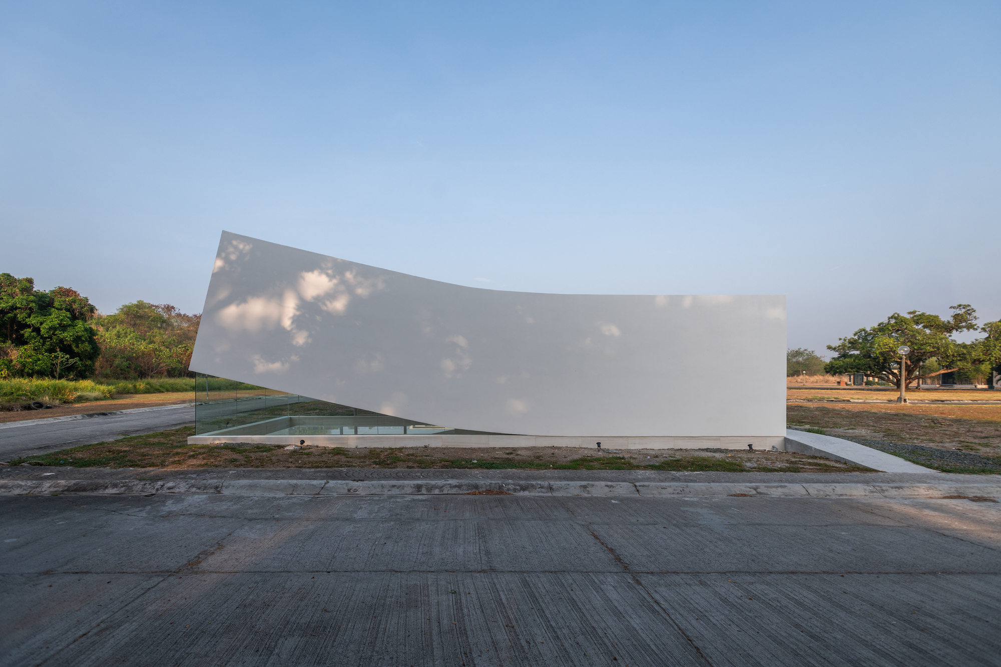 Street-level view of the white curved wall tilting over a glass base, with tree shadows dappling its surface at dusk