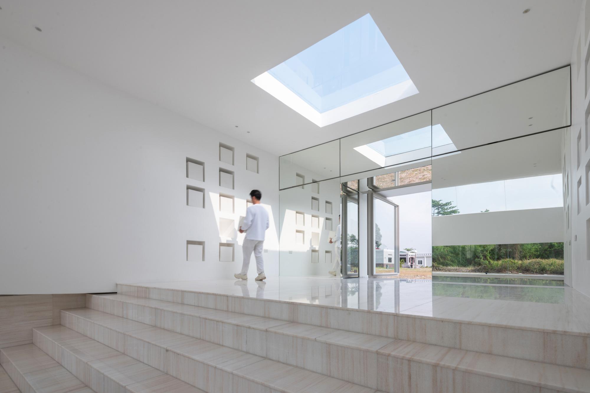 Person walking across the travertine platform toward a glass wall, beside a white wall with square niches and a skylight above