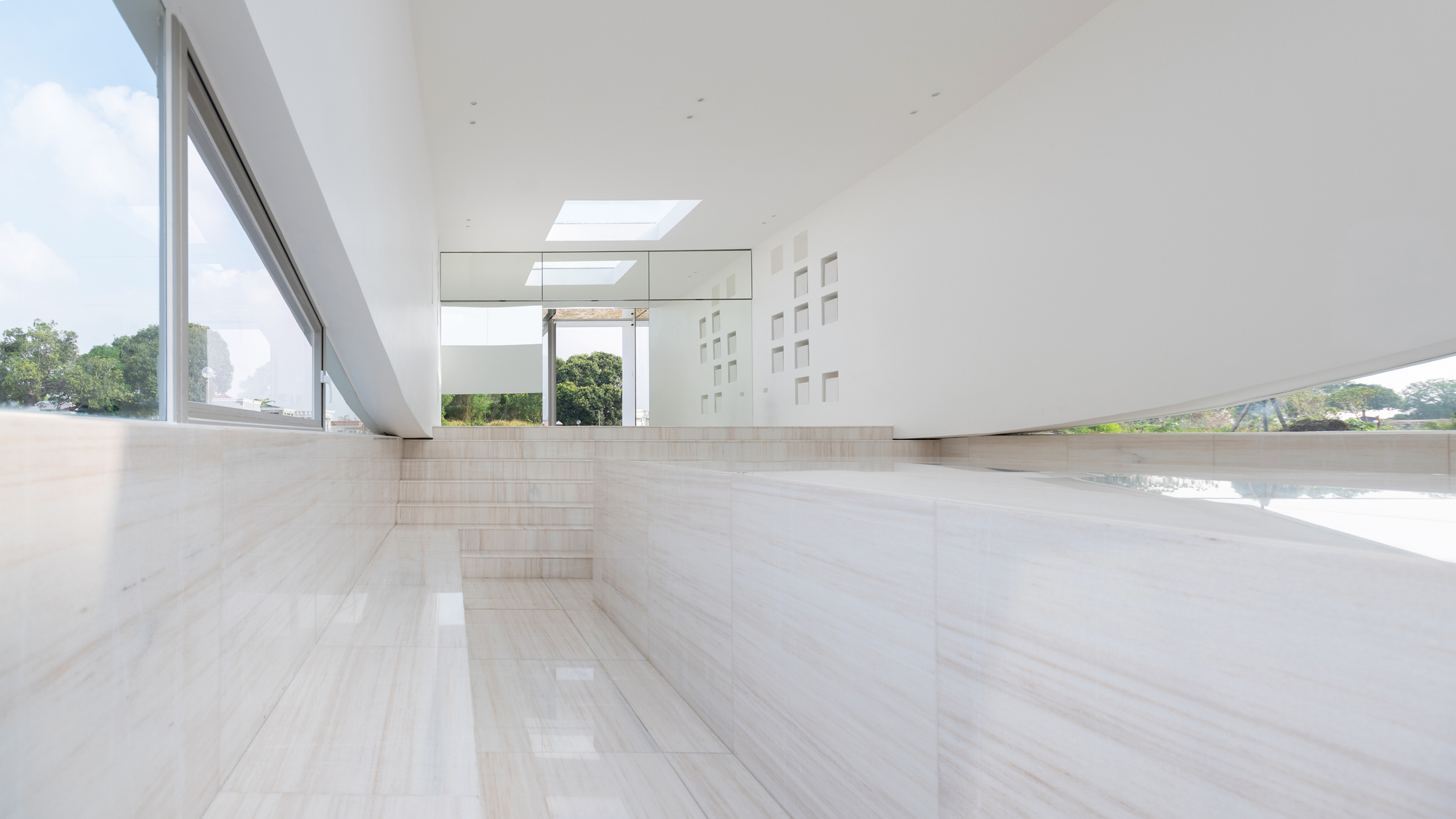 Long interior view of travertine steps and benches leading to a glazed wall, with a skylight and niche wall in daylight