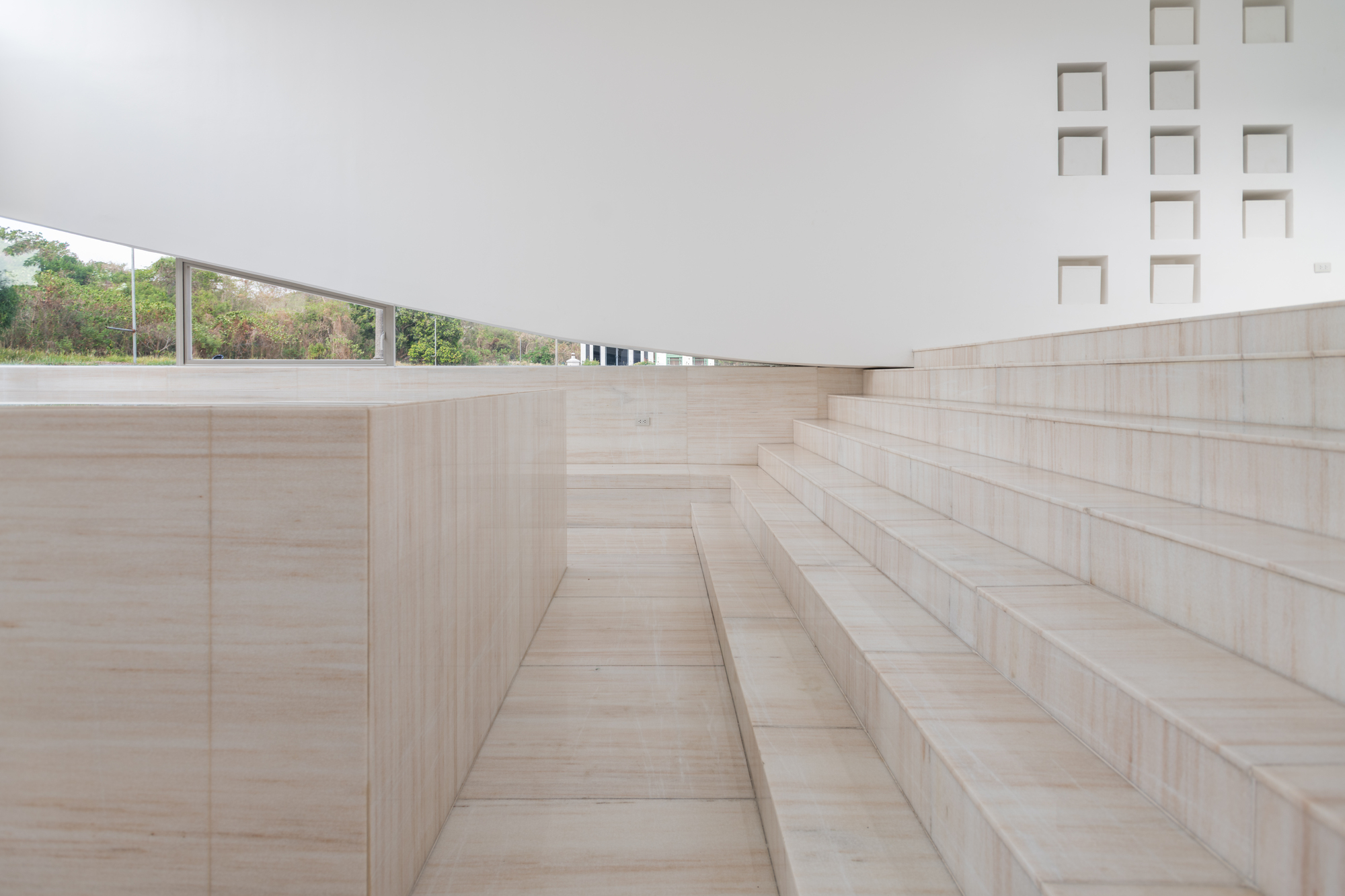 Pale travertine steps rising beside a stone bench block beneath a white wall with square niches, with green trees seen through a low window