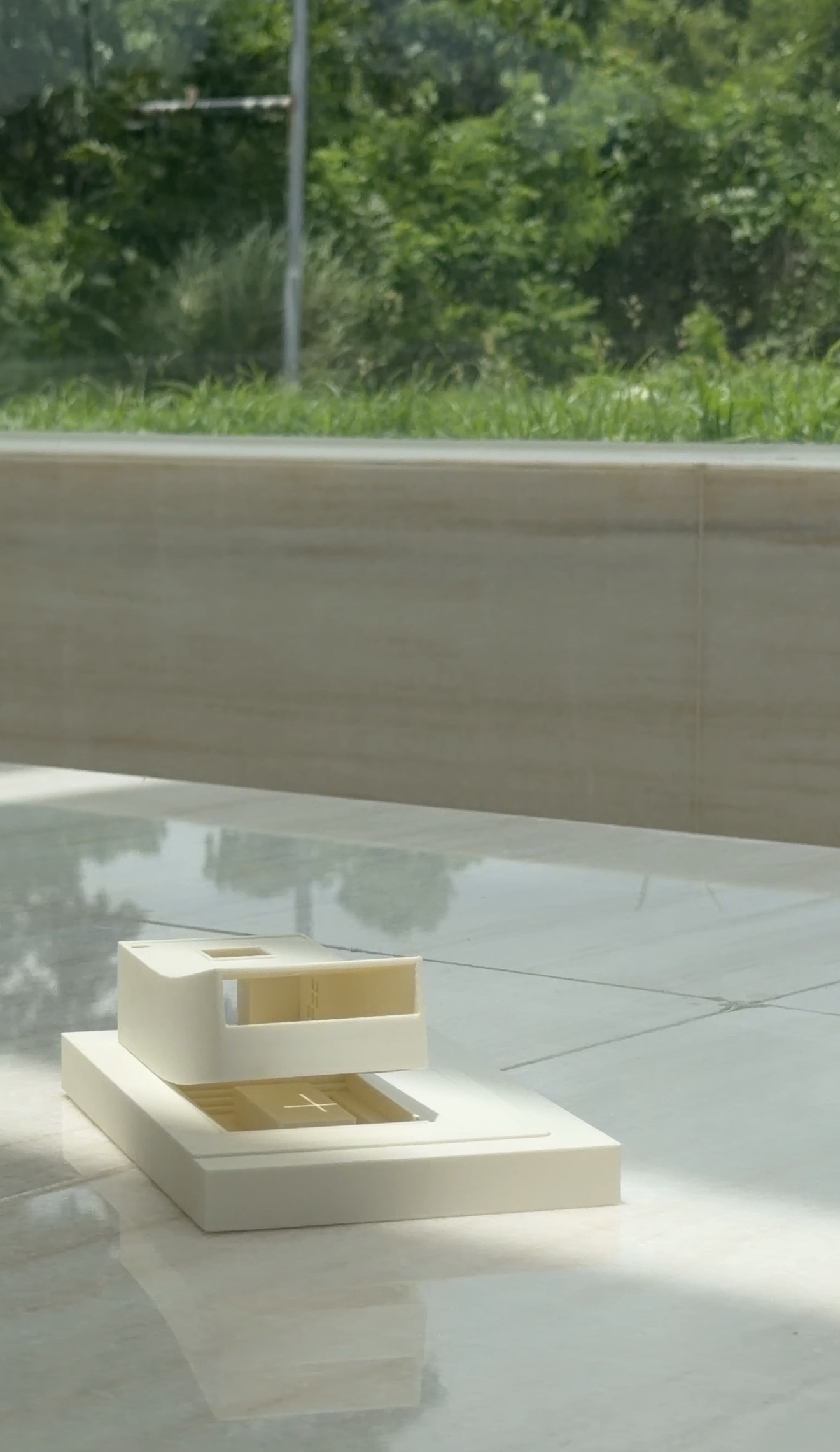 White scale model of a small building on a polished marble floor in patches of sunlight, with a stone wall and foliage behind