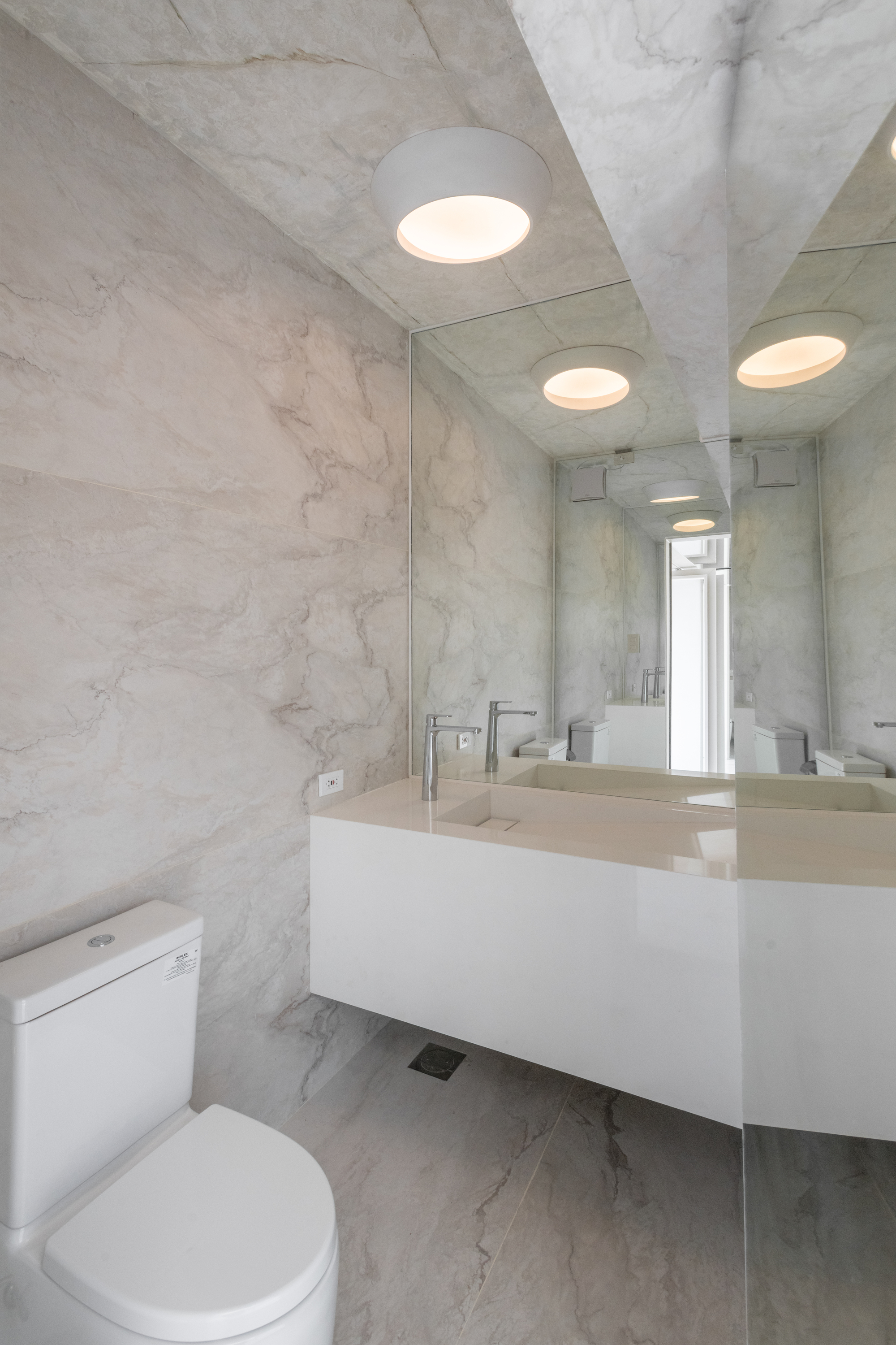 Powder room clad in veined marble slabs with a floating white basin, mirror wall and round ceiling lights