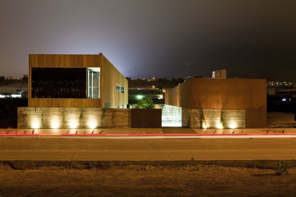 Street-level night view of timber-clad and rust-colored volumes on a board-formed concrete base, lit by ground uplights beside a light-streaked road