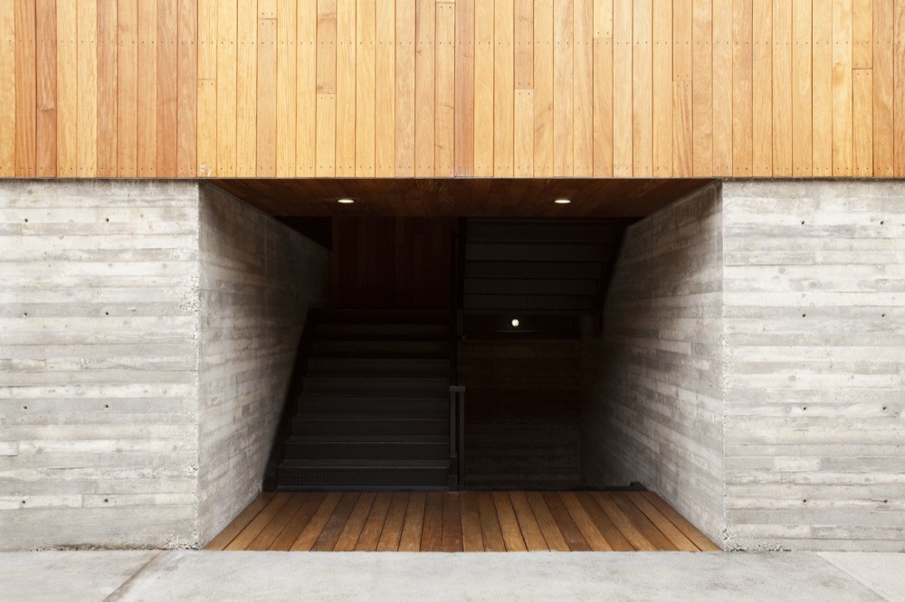 Recessed entry with board-formed concrete walls, a timber soffit and a dark stair descending, beneath vertical wood cladding in daylight