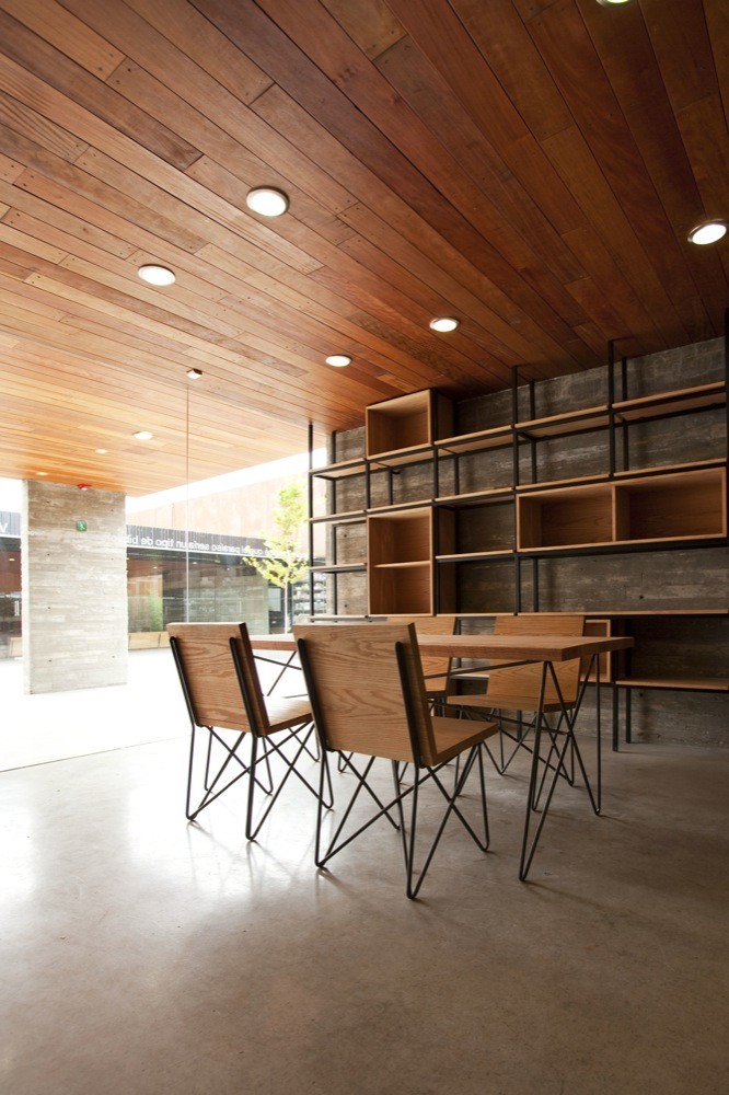 Dining table with wire-legged chairs before a steel and timber shelving wall, under a plank ceiling with round downlights
