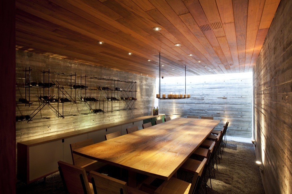 Long wine room with a timber table, bottle racks on a concrete wall and a candle chandelier near a bright skylit end