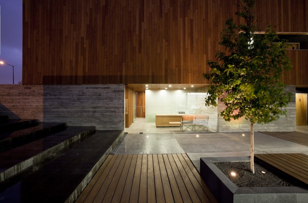 Entry court at dusk with a young tree in a stone planter, timber decking, black stone steps and a glazed lobby