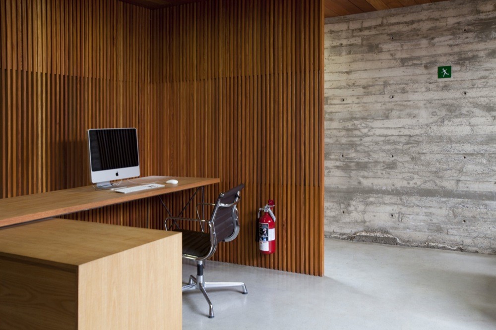Timber-slatted workstation wall with a desktop computer, office chair and fire extinguisher beside a board-formed concrete wall