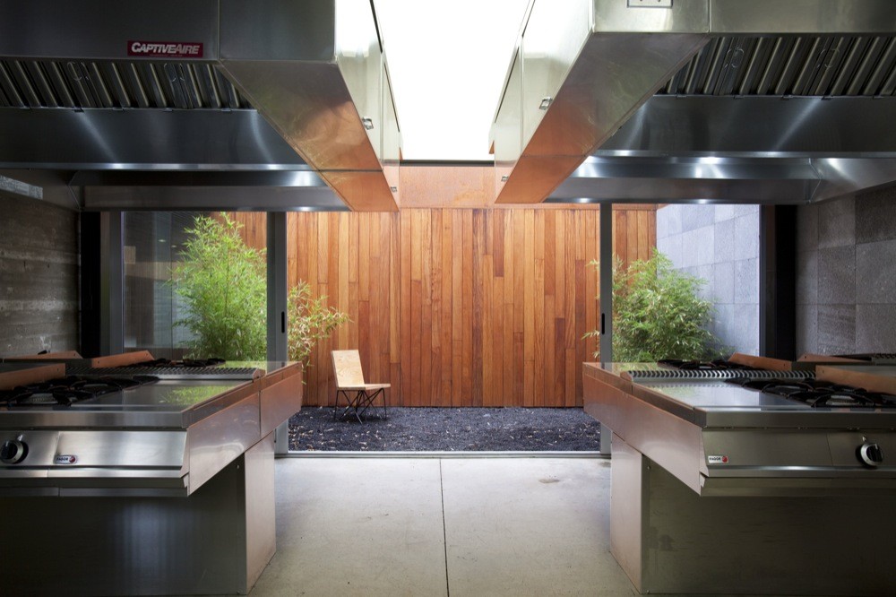 Two steel cooking islands under extraction hoods framing a timber-clad courtyard with bamboo, a wooden chair and black gravel